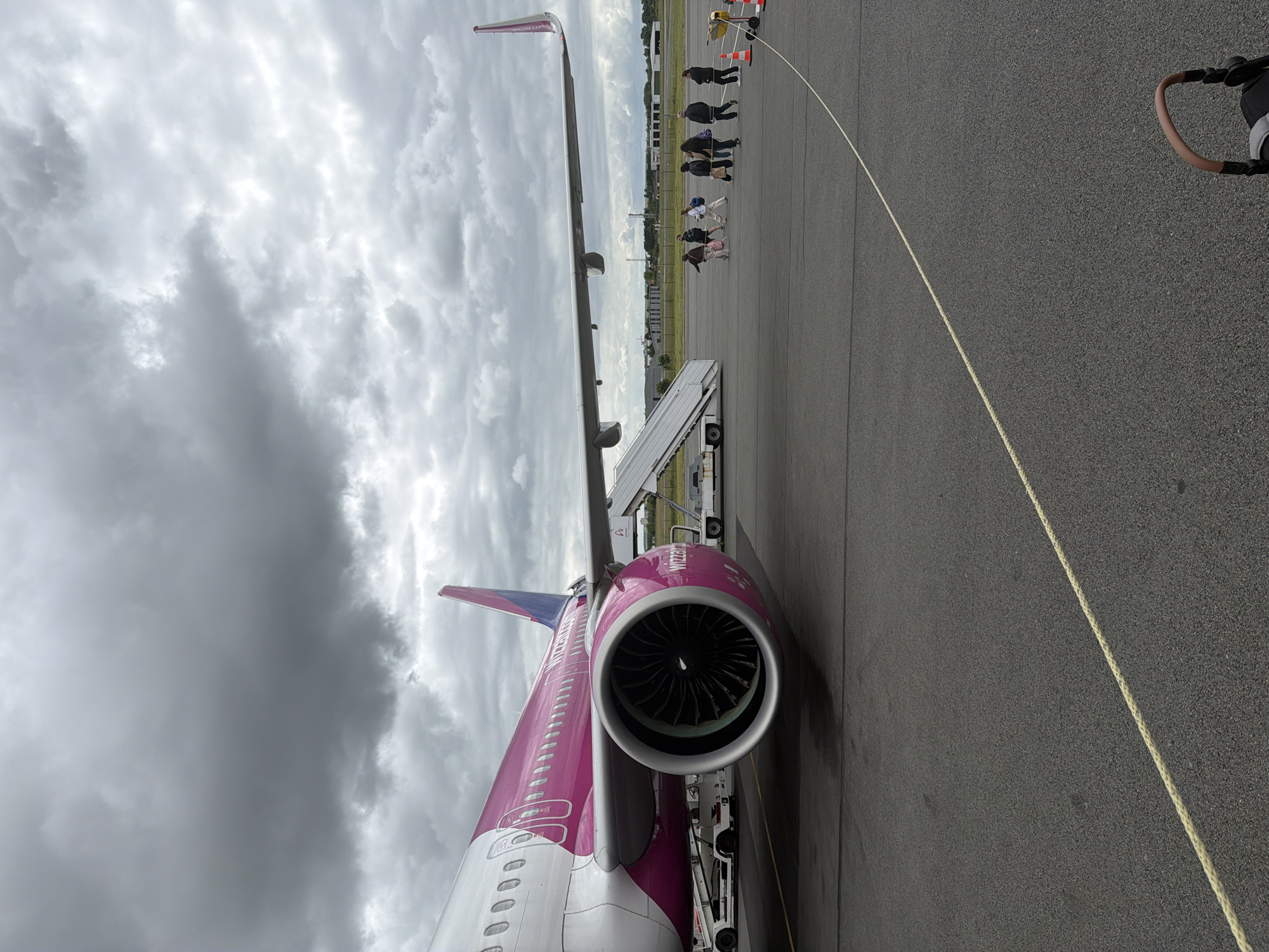 An overcast daytime scene at an airport tarmac in Tillé, France, shows a magenta and white Wizz Air aircraft. The image is taken from a low angle, looking upwards and along the side of the plane, which is rotated on its side.
The prominent subject is the large, single-aisle passenger jet, painted in Wizz Air's distinctive purple/magenta and white livery. The aircraft's right engine, with its visible fan blades, is in the foreground, and the fuselage extends towards the tail, featuring rows of windows. The right wing stretches into the distance, with a wingtip fence clearly visible. The text "WIZZAIR" is discernible on the side of the engine cowling, and "wizzair.com" can be vaguely made out on the wingtip.
To the right of the aircraft, several people are walking on the tarmac, appearing to be passengers or ground staff. They are heading towards or away from the aircraft, suggesting a boarding or disembarking process. A mobile boarding stair is positioned near the aircraft's entry door. In the far right foreground, part of a black stroller or pushchair handle is visible.
The ground is dark asphalt, with a light yellow line running diagonally across it, possibly indicating a safety boundary or taxiway edge. In the background, beyond the people, there are orange traffic cones and other airport equipment. The sky is heavily overcast with thick, grey clouds, indicating gloomy weather conditions.