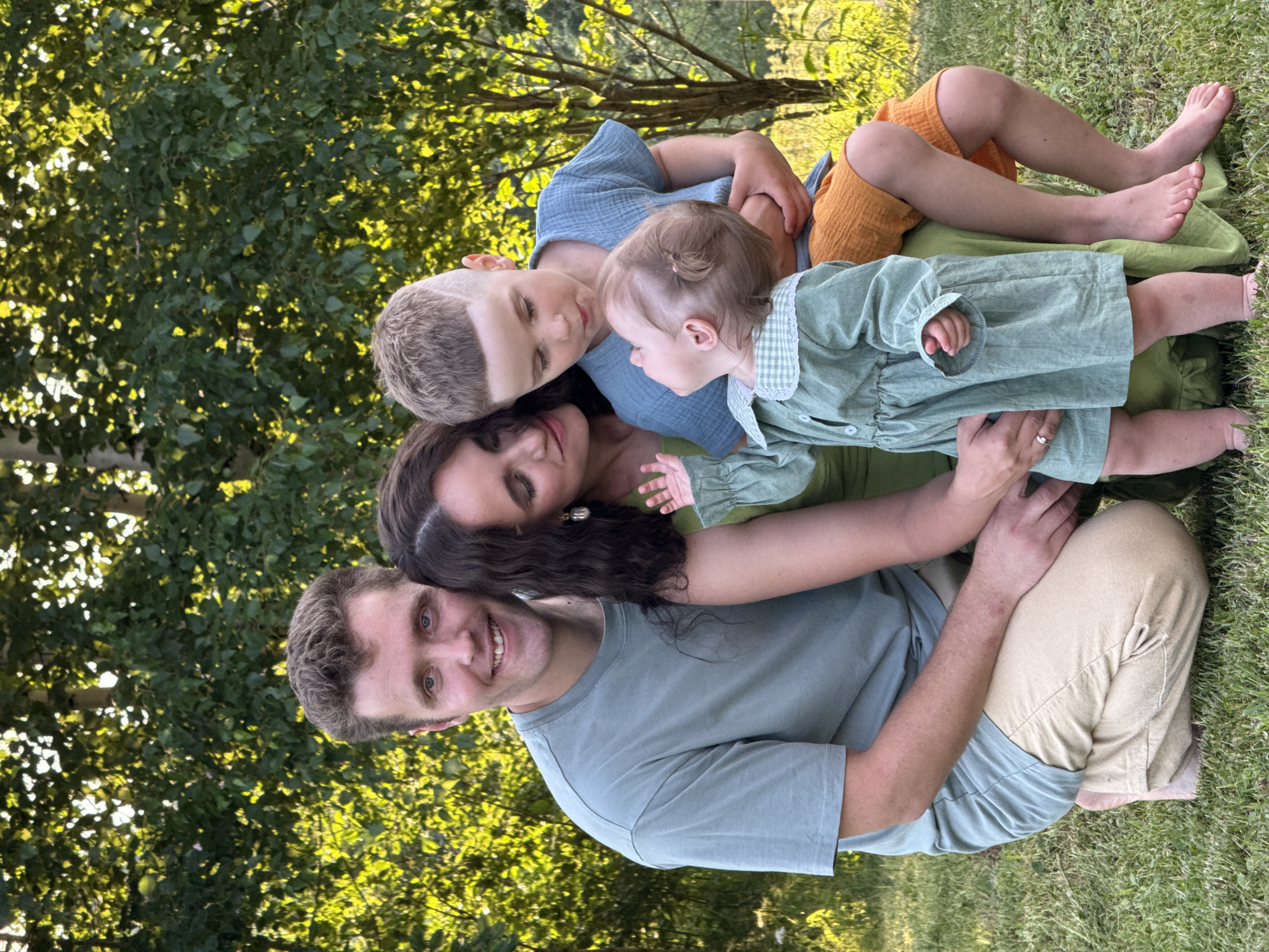 The image depicts a family of four posing for a photograph in an outdoor setting, likely a backyard or park, with lush green trees and foliage providing a natural backdrop. The sun is shining, casting dappled light through the leaves.
In the foreground, a man and a woman are positioned in a crouched stance. The man, on the left, wears a muted green t-shirt and tan pants. He is smiling directly at the camera. The woman, to his right, is dressed in a green dress. She is looking down towards the children.
Between the couple, two young children are visible. A boy, likely older, is seated on the woman's lap, wearing a blue t-shirt and orange shorts. He is looking down at a baby. The baby, a girl, is also in a green dress and is looking up at the boy. The family appears to be interacting affectionately, with the boy gently holding the baby.
The overall scene is one of a happy family moment, captured in a relaxed, natural environment. There are no specific identifiable landmarks to indicate the precise location within the city of Гаревая, Russia, other than the general description of an outdoor, green space.
