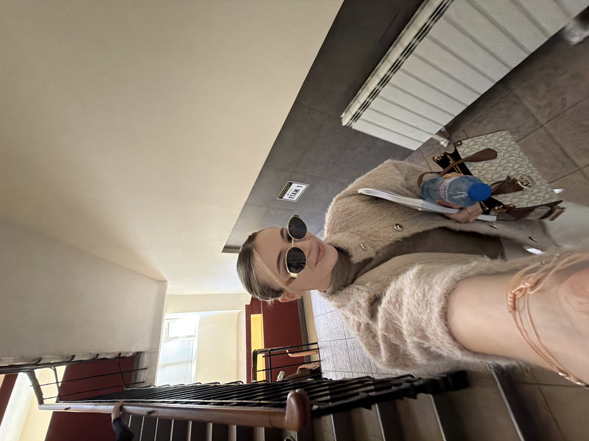 The image depicts a young woman with blonde hair, wearing sunglasses and a fuzzy cardigan, standing on a staircase. She is holding a water bottle and papers. The staircase has metal railings with dark brown wooden handrails. The background shows interior hallway features, including a radiator, a tiled floor, and a doorframe visible through an opening. A small sign reading "ETAK 1" is mounted on the wall. Based on the architectural style and the presence of Cyrillic script on the sign, the location is likely in Plovdiv, Bulgaria, but specific confirmation requires additional information.