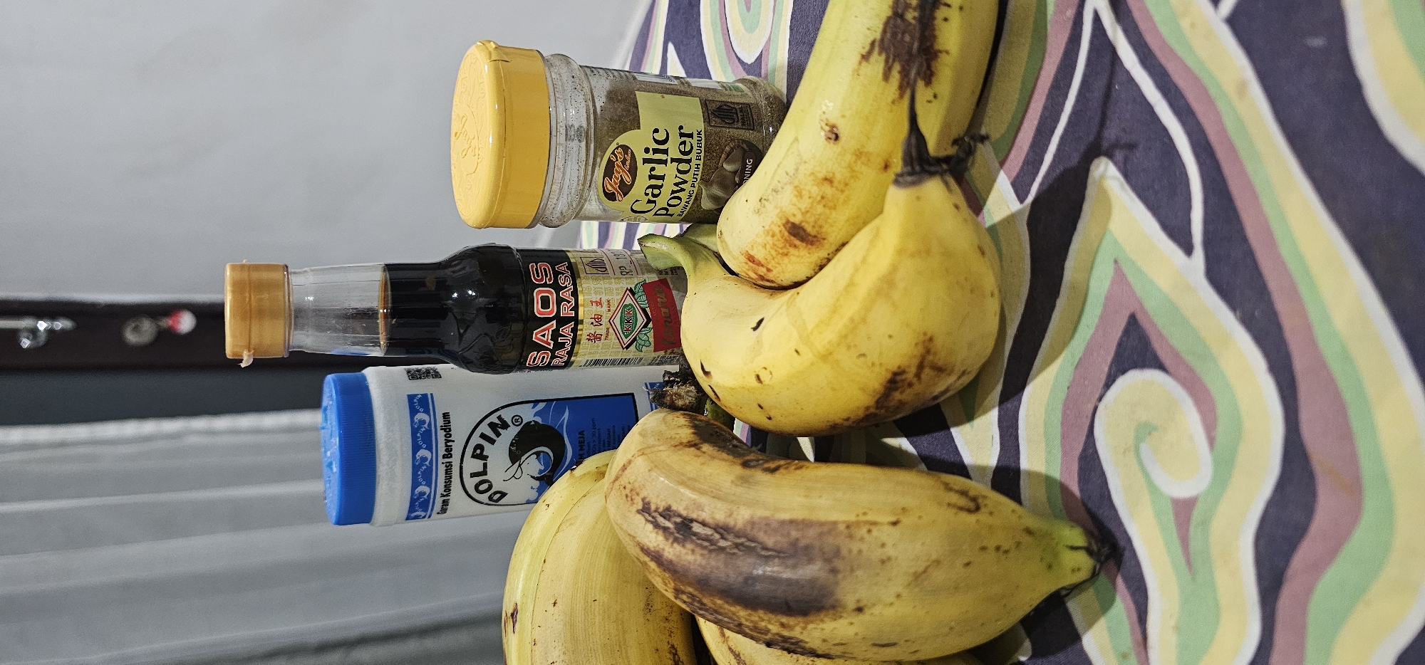 This media file presents a still life arrangement of food items and condiments, likely situated indoors on a table or counter in Denpasar, Indonesia. The image is oriented vertically.
The central subjects are a cluster of ripe bananas, several of which show dark brown spots indicative of their ripeness. Alongside the bananas, three common kitchen ingredients are visible. There is a clear bottle containing a dark brown liquid, identified by its label as "SAOS RAJA RASA" with Chinese characters "酱油王" (Soy Sauce King) and the "KIKK" brand logo. Adjacent to this is a clear plastic jar of "Jago's Garlic Powder," also labeled "BAWANG PUTIH BUBUK" (Indonesian for garlic powder), and "NING." The third condiment is a white plastic container with a blue cap and label, bearing the brand "GOLPIN" and stating "Garam Konsumsi Beryodium" (Iodized Consumption Salt), along with "MEJA" (table salt) and "MEGAH."
The items are resting on a surface covered with a distinctive fabric or material featuring a swirling, striped pattern in light green, yellow, pink, and dark blue or purple hues. The background is out of focus, showing a light grey wall or surface with a horizontal dark element and some small, indistinct metallic objects. No specific activity or event is taking place, and no people or explicit emotions are visible. The lighting suggests an indoor daytime setting.