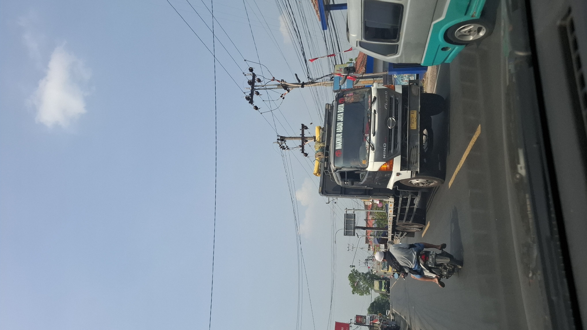 The image captures a street scene in Cikarang, Indonesia, under a clear blue sky with sparse white clouds. A large Hino truck with "MAKMUR ABADI JAYA RAYA" printed on its windshield is the central focus, driving on an asphalt road. The truck is black and white, with yellow protective coverings on its roof rack. To the left of the truck, a motorcycle with two occupants, one driving and one passenger, is traveling in the same direction. The driver wears a white cap and a light gray shirt, while the passenger has on a light gray shirt and blue shorts. A teal and silver van is visible to the right of the truck. Electrical poles with numerous wires line the sides of the road, and various buildings, including shops, can be seen in the background. The road is marked with yellow lines. The photograph appears to be taken from inside a vehicle, with the dashboard visible in the foreground.