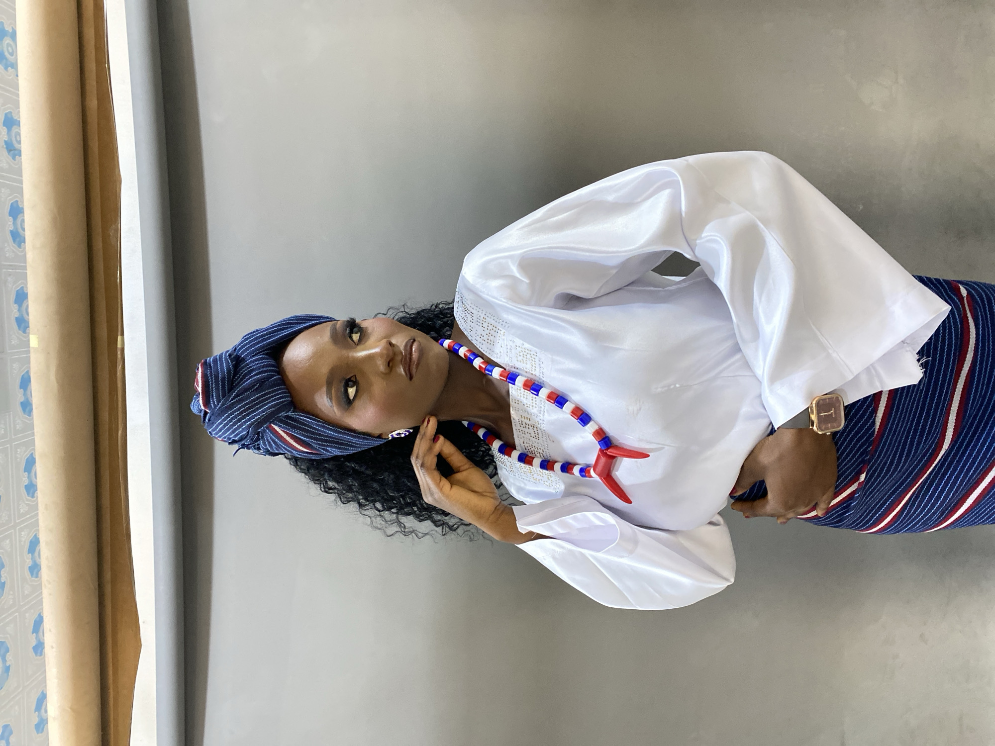 In Gura Top, Nigeria, a Black woman with dark, curly hair, adorned with a blue, white, and red patterned headwrap, poses against a grey backdrop. She wears a white, long-sleeved satin top embellished with rhinestones around the neckline, and a colorful beaded necklace featuring red, white, and blue beads, with a red pendant. Her attire is completed by a blue and white striped skirt. She is positioned facing right, with her left hand touching her chin and her right hand resting on her waist. A gold-cased wristwatch is visible on her right wrist. The environment appears to be indoors, with a rolled-up beige material and a patterned ceiling visible at the top of the frame.
