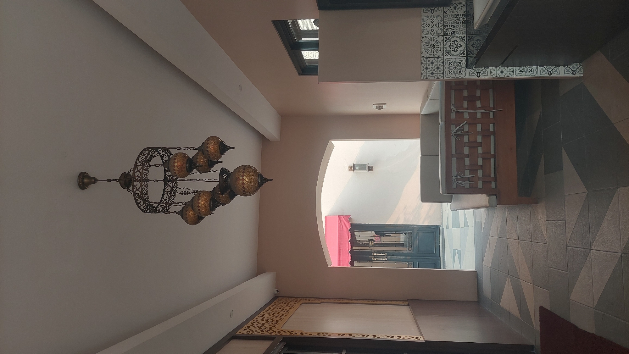 The image depicts an indoor scene, likely a cafe or waiting area, with a low-angle perspective. A large, ornate chandelier with multiple amber-colored glass globes hangs from the ceiling. The walls are painted a soft peach color. To the left, there is a decorative wall panel with a gold geometric pattern. Further left, a dark door is partially visible.
In the center of the image, an arched entryway leads to an outdoor area with a red awning and a dark double door. The floor is tiled with a geometric pattern in shades of gray and beige.
A light-colored sectional sofa with wooden accents is positioned in the foreground, facing the arched entryway. To the right of the sofa, a section of a dark countertop with a white sink is visible, adorned with black and white patterned tiles. A window with a dark frame is above the countertop.
The overall impression is one of a well-decorated and comfortable space. The lighting suggests it might be late afternoon or early morning, given the soft, diffused light. The image does not contain any identifiable people or specific actions. There is no information within the image to suggest the location is Cileungsi, Indonesia.