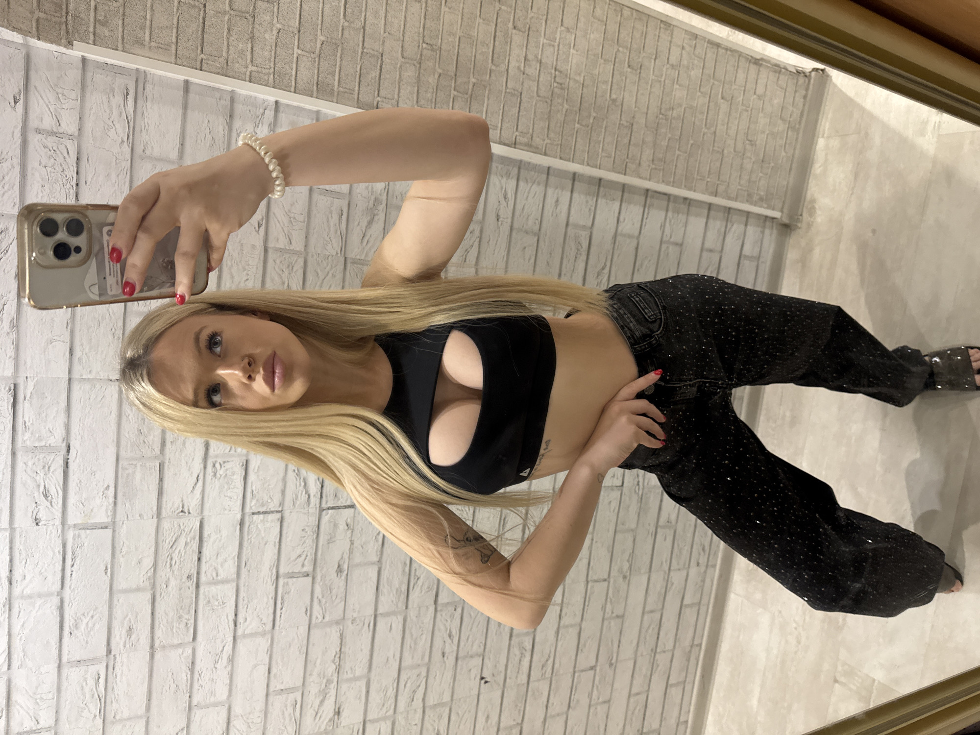 A young woman with long blonde hair is taking a mirror selfie. She is wearing a black crop top with a cutout design and dark, sparkling jeans. On her left wrist, she has a pearl bracelet. Her right hand is holding a gold-colored phone. The background is a white brick wall, suggesting an indoor setting. The lighting appears to be artificial, and the overall mood is casual. The photograph is oriented vertically.