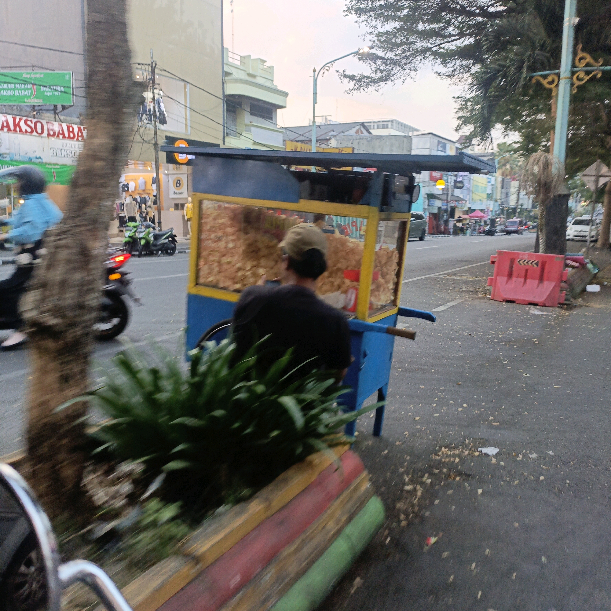 The image depicts a street scene in Salatiga, Indonesia, likely captured during the late afternoon or early evening, indicated by the soft lighting and the gentle sky. In the foreground, a portion of a vehicle, possibly a motorcycle given the visible chrome handlebars and a sliver of a tire, is captured in motion, creating a blurred effect. To the left, a textured tree trunk is prominently featured, partially obscuring the background.
Adjacent to the tree is a food cart, its blue base and yellow frame contrasting with the dark asphalt of the road. The cart is filled with a large quantity of what appears to be fried snacks, possibly crackers or chips, displayed behind glass. A person, seen from the back, wearing a black shirt and a light-colored cap, is tending to the cart, their posture suggesting they are either serving or preparing food.
Beyond the food cart, the road stretches into the distance, lined with various buildings that suggest a commercial or urban area. Signs for businesses, including "AKSO BABAT" and "BAKSO BARAT," are visible on the buildings, indicating food-related establishments. The street is active with traffic, including motorcycles and cars, some of which are blurred due to motion. Red traffic barriers are placed on the side of the road, suggesting a possible construction or temporary lane closure. Green plants are also visible, adding a touch of nature to the urban landscape. The overall impression is that of a busy, everyday street in a city in Indonesia.