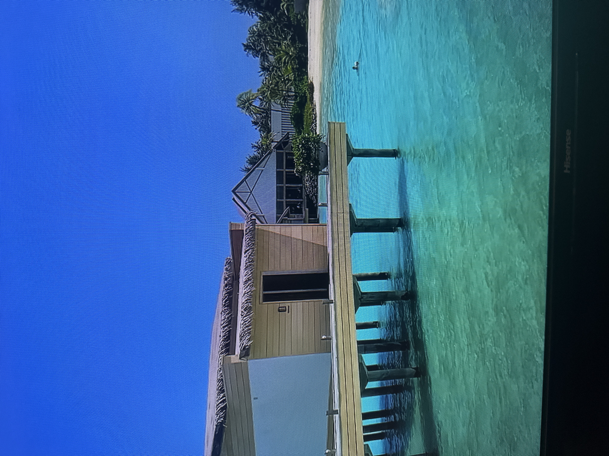 The image depicts a tropical resort scene with overwater bungalows and a clear turquoise ocean under a bright blue sky. A person is visible swimming in the water. In the background, there is a sandy beach lined with palm trees and a large building with a distinctive architectural style. The scene is calm and serene, evoking a sense of vacation and relaxation. The location is not in Kyiv, Ukraine.
