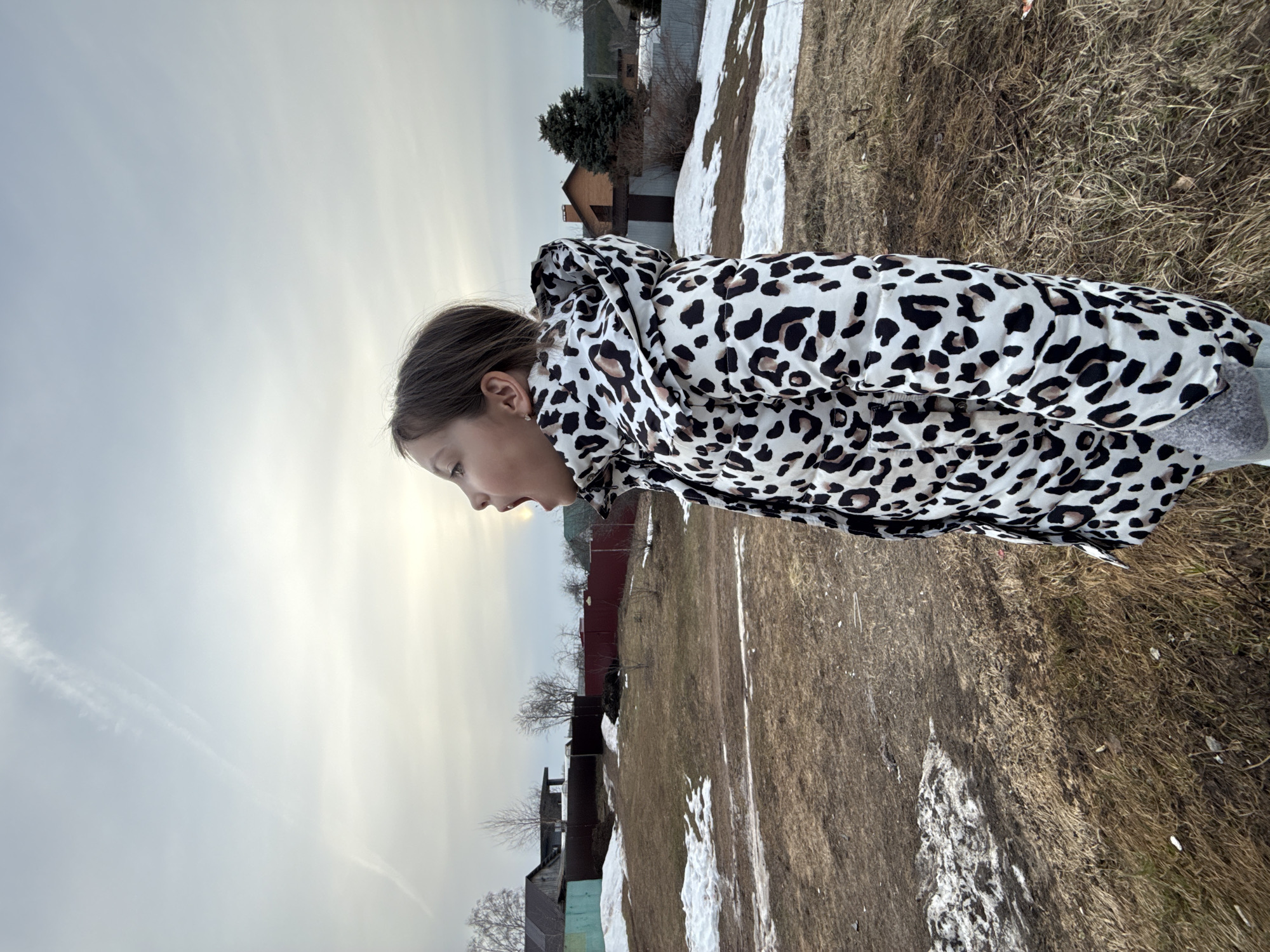 A young girl stands in a grassy field with patches of snow in Votkinsk, Russia. She is wearing a white and black leopard-print jacket with the hood up. Her head is angled down. Behind her are houses with metal roofs and trees with bare branches. The sky is overcast with thin, streaky clouds. The ground is covered in a mixture of dry grass, dirt, and melting snow.