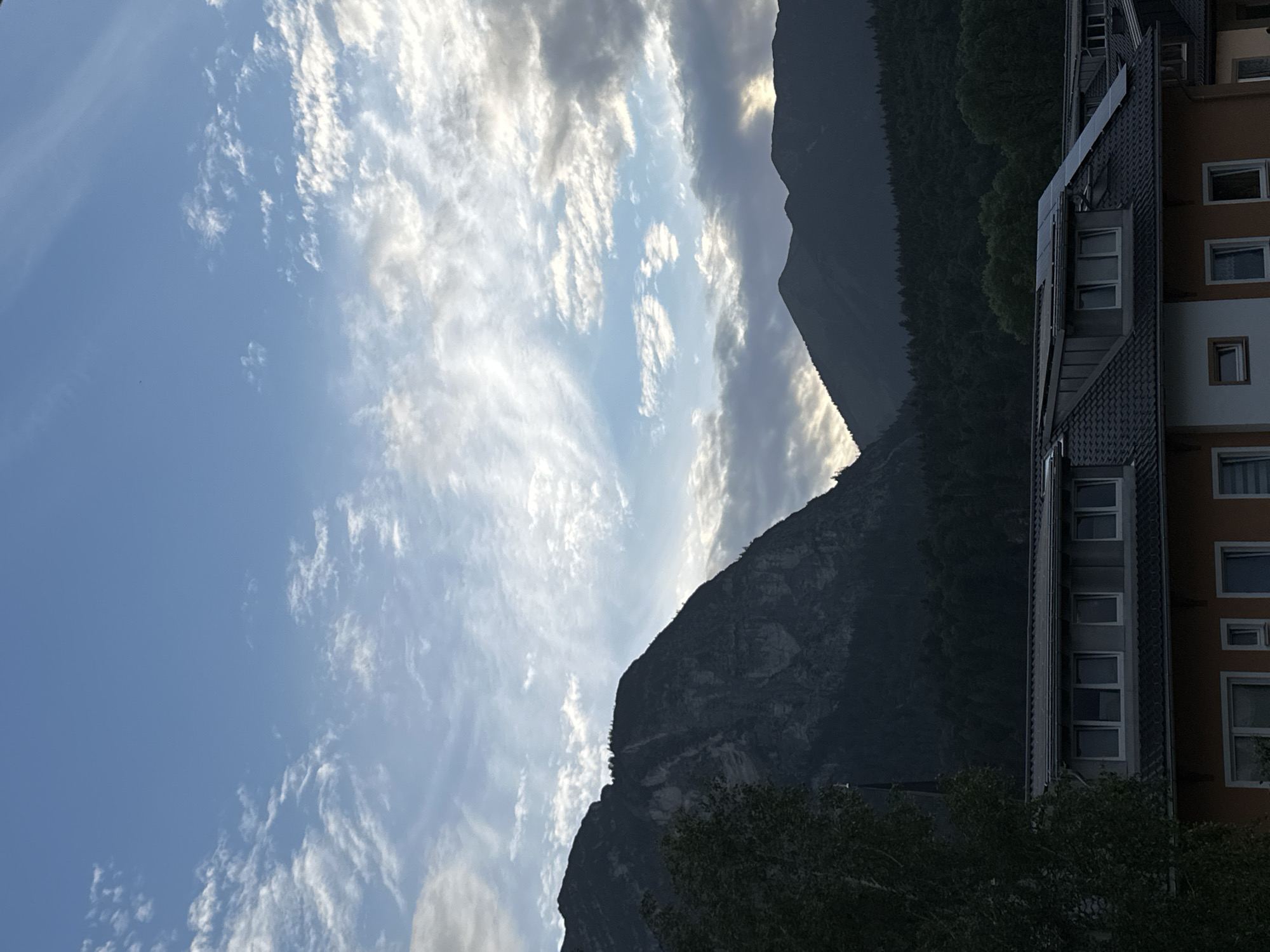 The image depicts a mountainous landscape with a building in the foreground, likely taken from an elevated position within the city of Garmisch-Partenkirchen, Germany. The sky is partly cloudy with a mix of blue sky and wispy white clouds, with hints of sunlight breaking through on the right side. Dominating the mid-ground are several dark, heavily forested mountains, with a prominent, jagged peak on the left. In the foreground, the roof of a building is visible, featuring dark shingles and several solar panels. The building itself appears to be multi-story, with visible windows and an orange-colored facade on the lower level. A large tree with dark green foliage partially obstructs the view on the left side of the image. There are no people or animals visible. The overall impression is a serene, natural scene juxtaposed with modern infrastructure.
