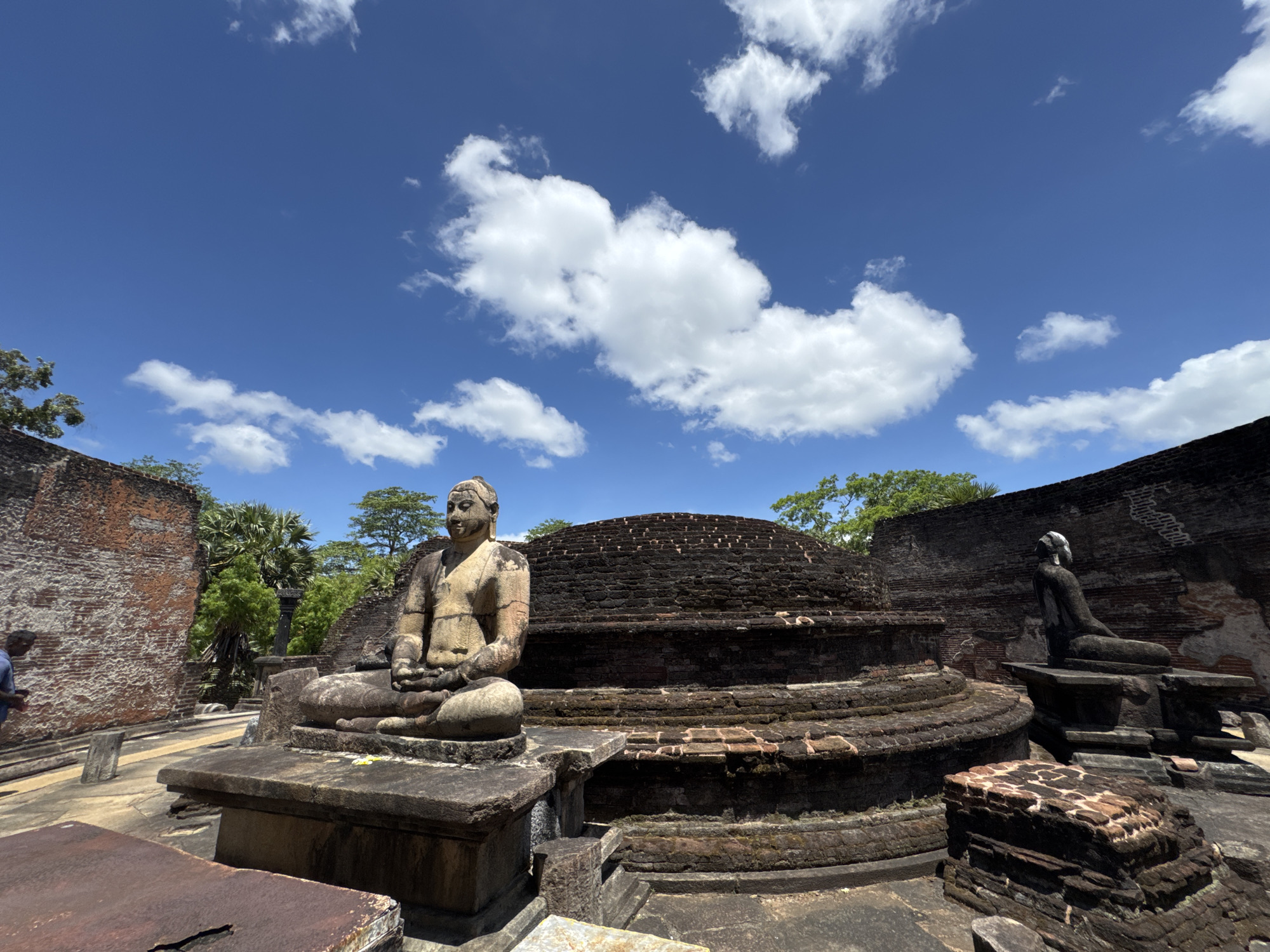 The image captures a scene from Polonnaruwa, Sri Lanka, featuring ancient ruins under a bright blue sky with scattered white clouds. In the foreground, a weathered stone statue of a seated Buddha is prominently displayed, its details worn by time. Behind the statue is a stepped, brick structure, likely a part of the historical site. To the right, another seated statue is visible, accompanied by additional brick ruins. The environment consists of aged brick structures and stonework, indicative of the site's historical significance and state of preservation.