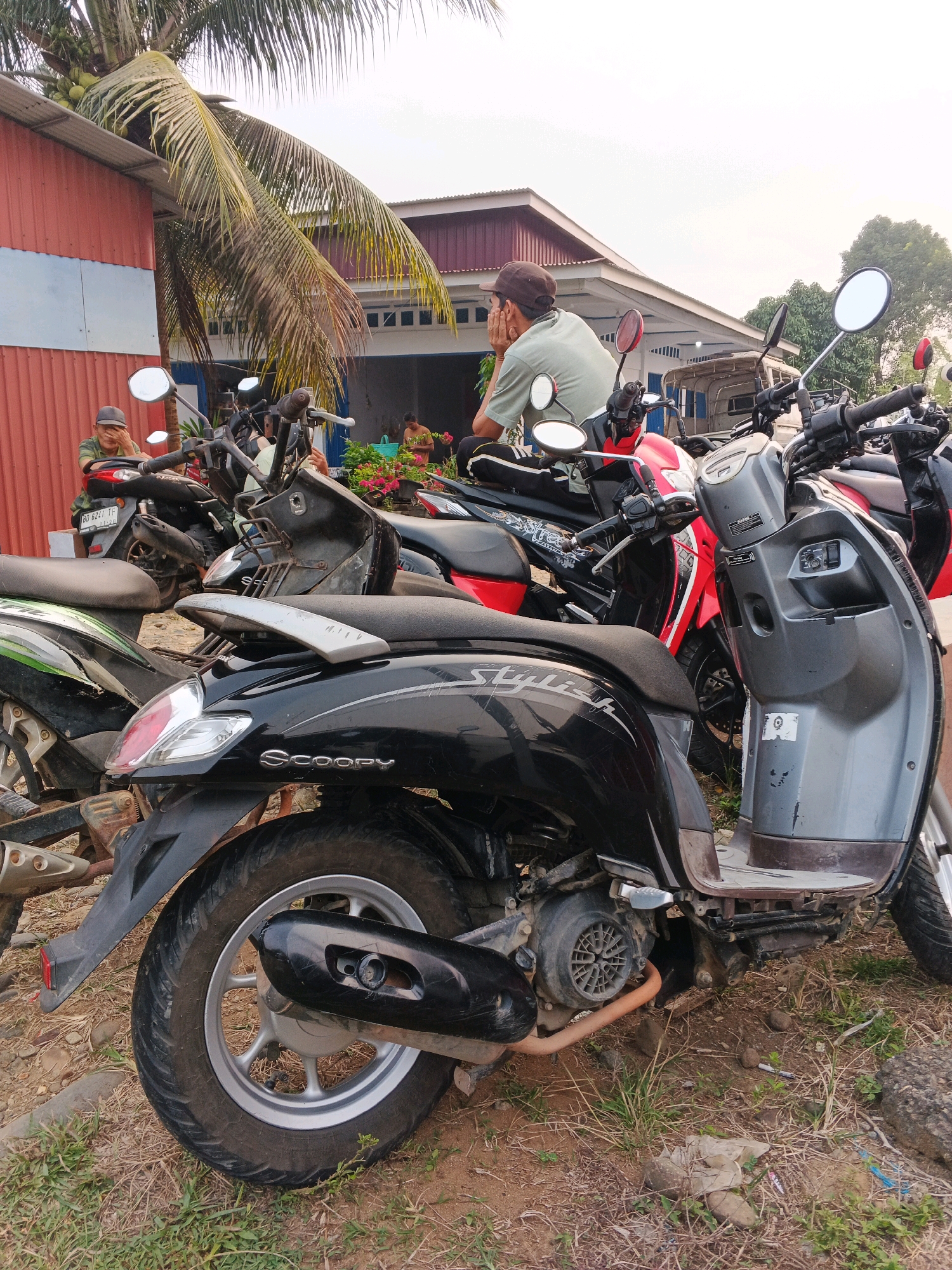 In Brangan Mulya, Indonesia, several parked motorcycles are visible in front of a building. A man wearing a cap and a light green t-shirt is seated on one of the motorcycles, resting his head in his hands. In the background, two other individuals are standing near the building. The environment appears to be a roadside or an open area with dry grass and dirt. A coconut tree with coconuts is leaning over a red-colored structure. The sky is overcast.
