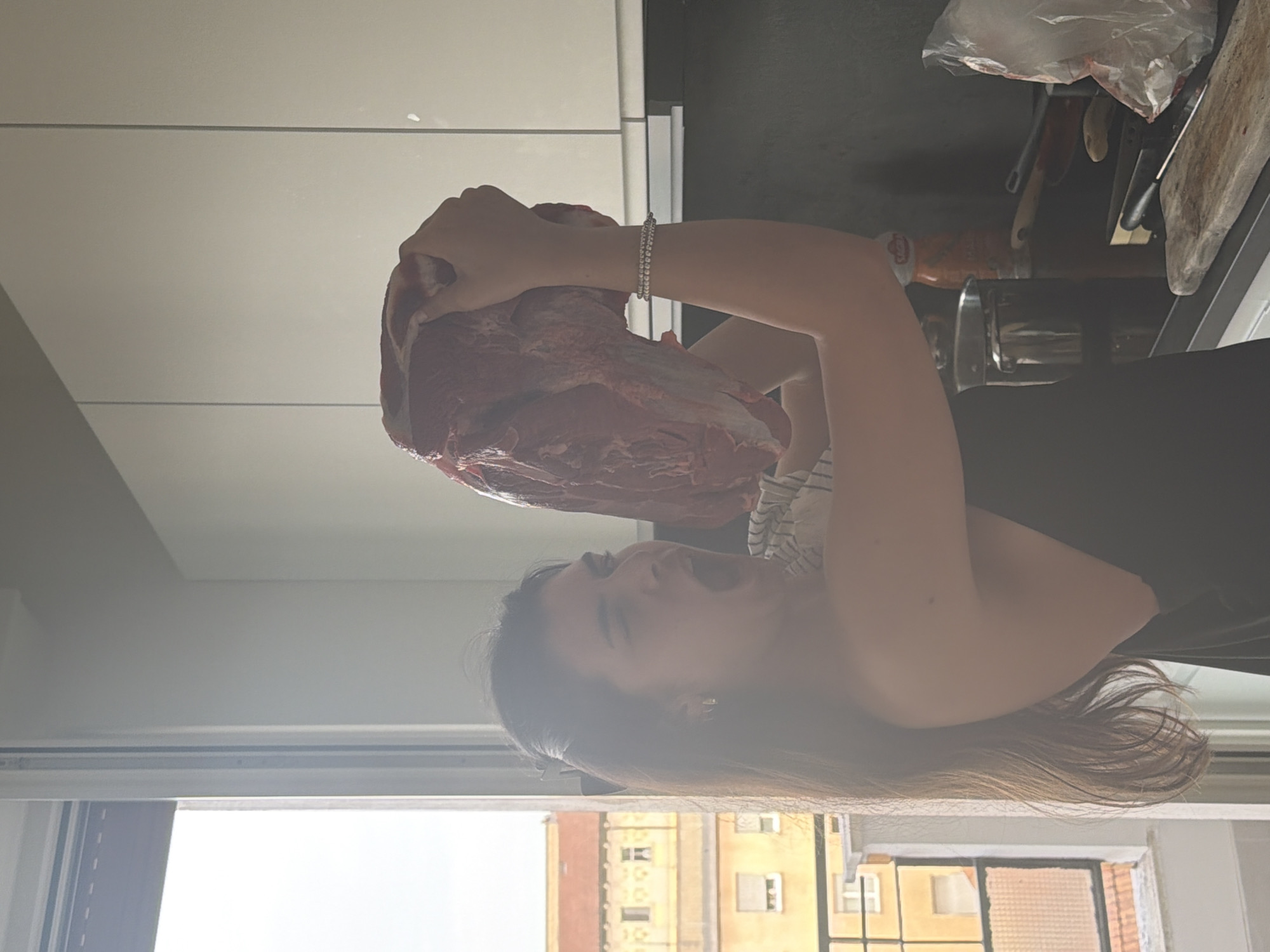 A young woman in Milan, Italy, holds a large piece of raw meat. She is standing in a kitchen with a window overlooking a building with multiple windows and apartment-style dwellings. She is smiling with her mouth open, as if excited about the meat. She is wearing a striped shirt and a bracelet. The kitchen counter behind her contains cooking utensils, including knives and a cutting board.