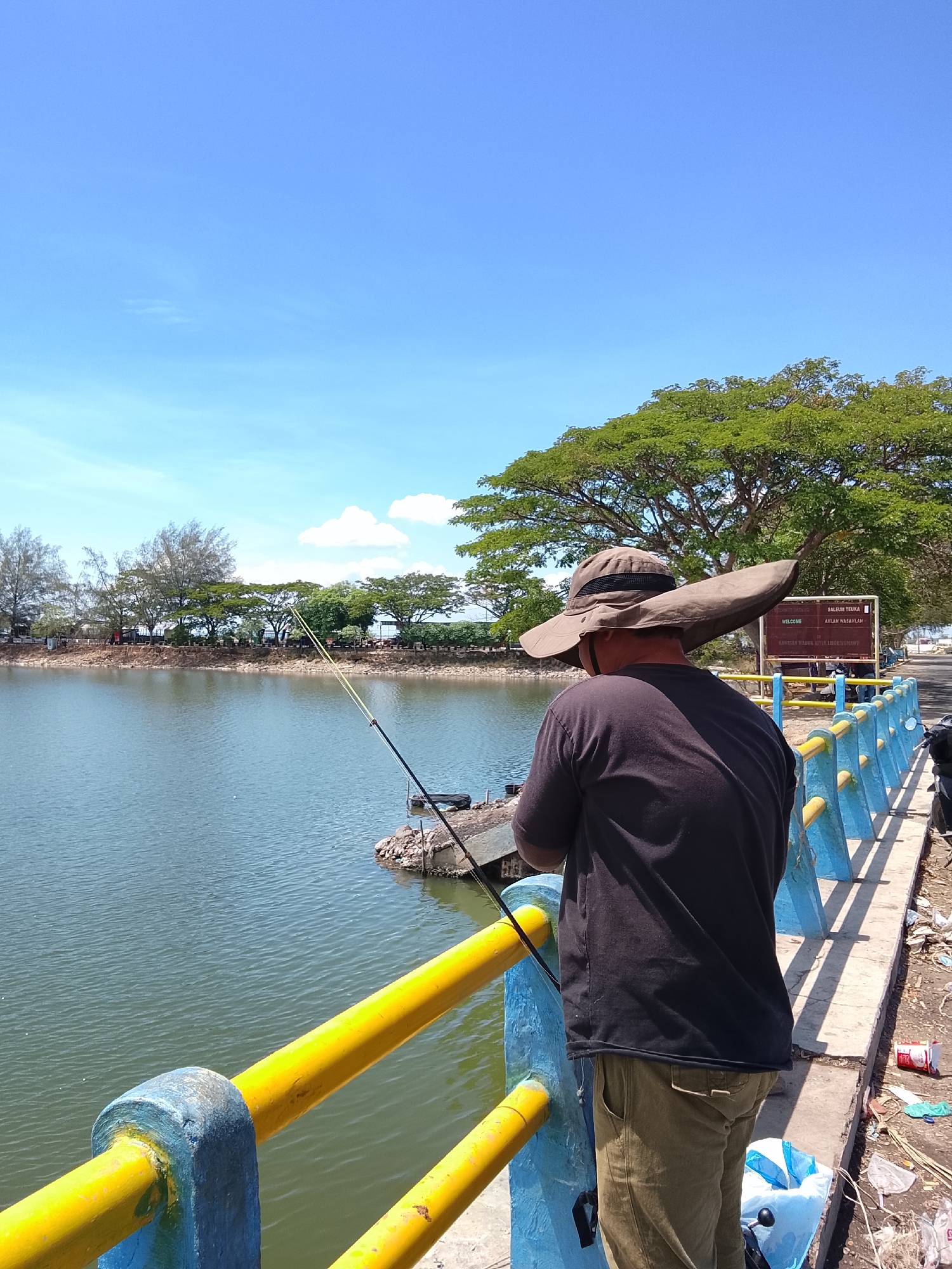 A lone fisherman, wearing a wide-brimmed hat and a dark t-shirt, stands on a concrete embankment, casting a fishing rod into a calm body of water. The embankment is bordered by a blue and yellow railing, with debris scattered along the ground. In the background, a line of trees separates the water from a road, with a signpost visible in the distance. The sky is a clear blue with scattered white clouds. The location is likely the reservoir area of the Arun Lhokseumawe Gas Project in Lhokseumawe, Aceh, Indonesia, a common spot for recreational fishing.
