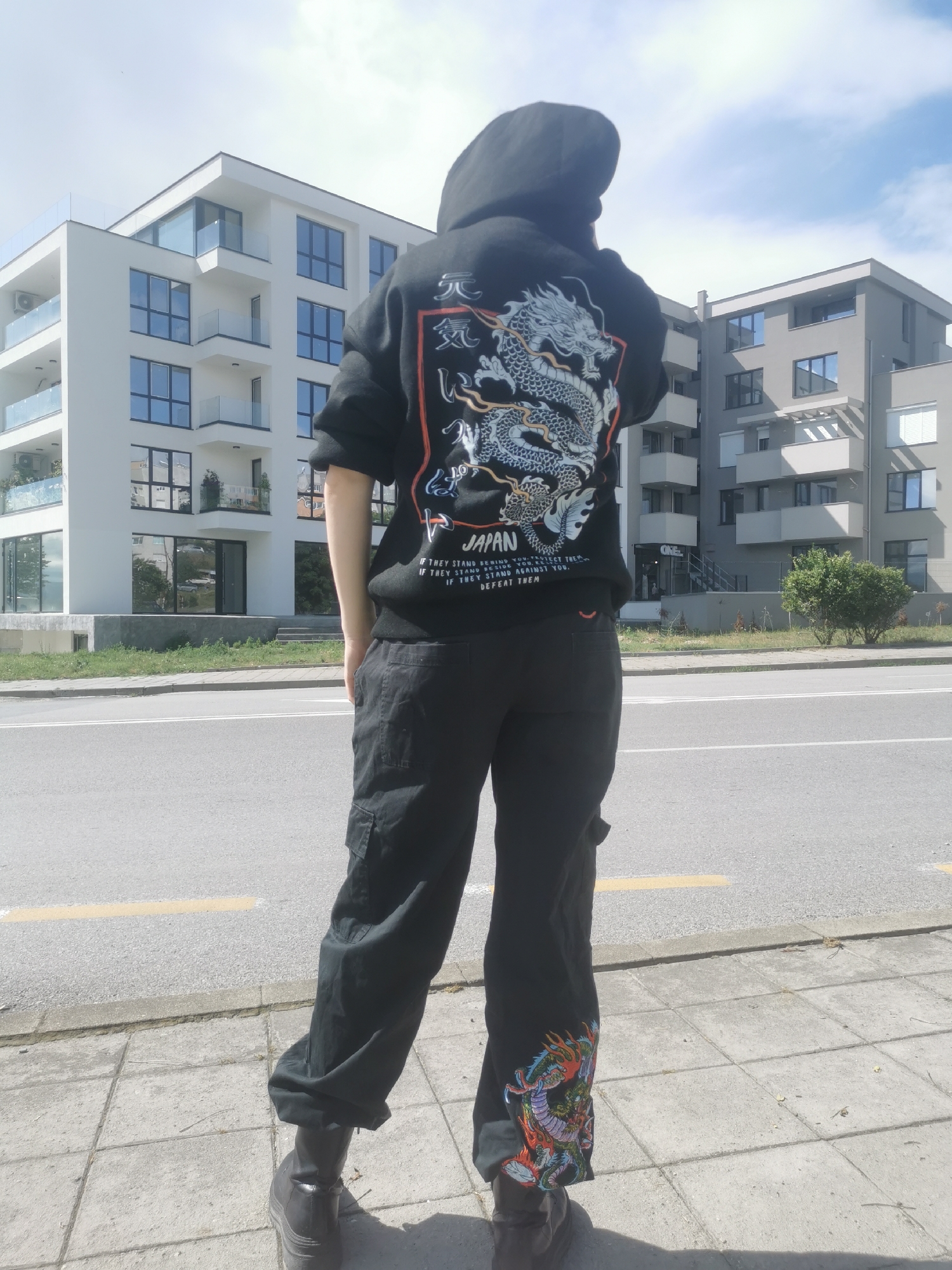 A person is captured from behind, standing on a sidewalk next to a road in Blagoevgrad, Bulgaria, on what appears to be a bright, sunny day with a partly cloudy sky. The individual is wearing a black hooded sweatshirt with an elaborate white and blue dragon design on the back, highlighted with red and orange accents. Above the dragon, Japanese characters "元 氣" (Genki) are visible, and below it, the word "JAPAN" is printed. Further English text reads: "IF THEY STAND BEHIND YOU PROTECT THEM. IF THEY STAND BESIDE YOU RESPECT THEM. IF THEY STAND AGAINST YOU. DEFEAT THEM". The person is also wearing black cargo pants, with a vibrant, multi-colored dragon design (featuring red, orange, blue, and green) embroidered near the cuff of the right leg. Dark, chunky boots complete the outfit.
In the background, two modern multi-story apartment buildings are visible. The one on the left is white with numerous dark-framed windows and balconies featuring clear glass railings and air conditioning units. The building on the right is gray, with similar windows and balconies. The road in front of the sidewalk has painted white and yellow lines, and sparse green vegetation, including a small bush, grows between the road and the buildings. A commercial sign with the word "ONE" can be seen on the ground floor of the gray building. The scene depicts a casual urban setting.