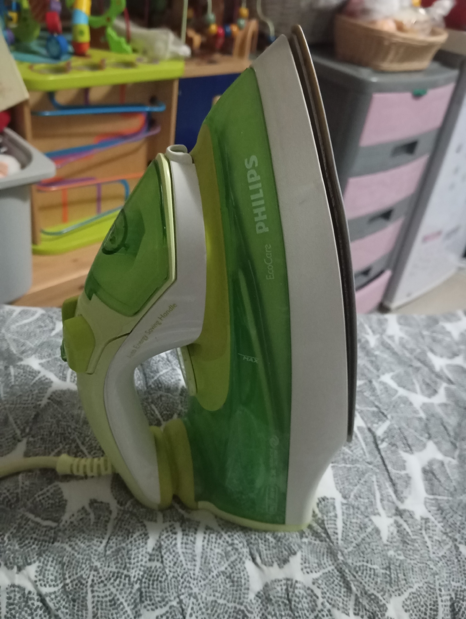 A green and white Philips EcoCare steam iron stands upright on its heel rest in an indoor domestic setting. The iron features a translucent green water tank with "PHILIPS" and "EcoCare" branded vertically and horizontally, respectively, and a "MAX" fill line indicator. The handle is white and light green, with text "Auto Energy Saving Handle" visible. A light yellow power cord with a coiled strain relief extends from the base. The iron rests on a patterned fabric surface, dark grey, and white, resembling stylized leaf or organic motifs.
In the blurred background to the left, a multi-level wooden shelf or storage unit contains various colorful children's toys, including sections of blue, orange, and purple plastic tracks. A grey container is partially visible on the far left. To the right, a blurred dark grey plastic storage unit with three pink-fronted drawers is present, topped by a woven basket containing light-colored, soft items. No individuals are present, and no specific actions are being performed. The environment appears to be a living space, possibly a child's room or utility area. The exact location in the city of Dubai, United Arab Emirates, cannot be determined from the visual content of the image.