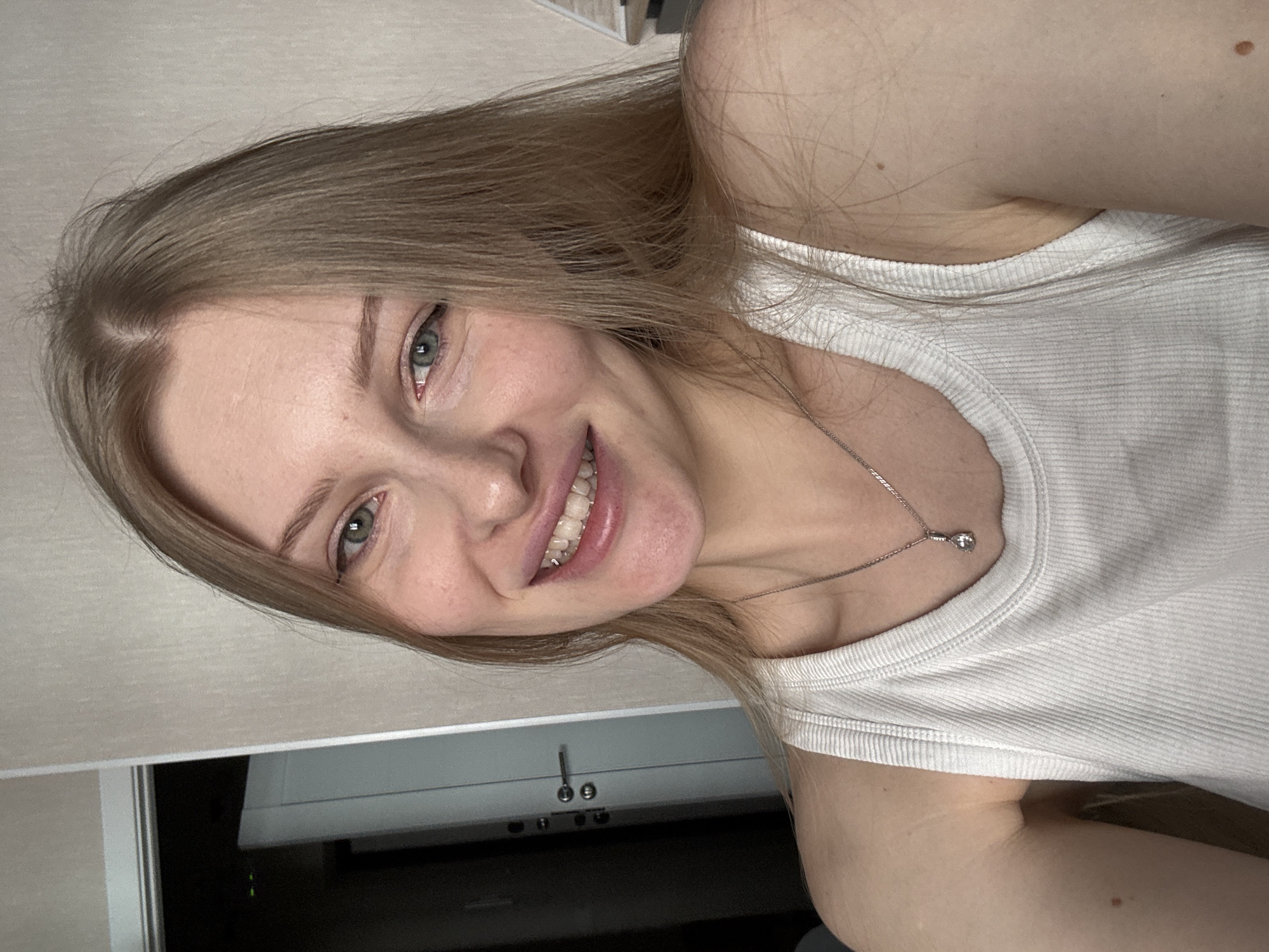 A selfie of a fair-skinned young woman with blonde hair and blue-grey eyes. She is smiling, revealing teeth with braces. She is wearing a white tank top and a silver necklace with a pendant. The background is blurry, but a grey cabinet with silver handles and what appears to be a dark room can be seen. The location of the image cannot be confidently determined to be in Perm, Russia, or any other location.