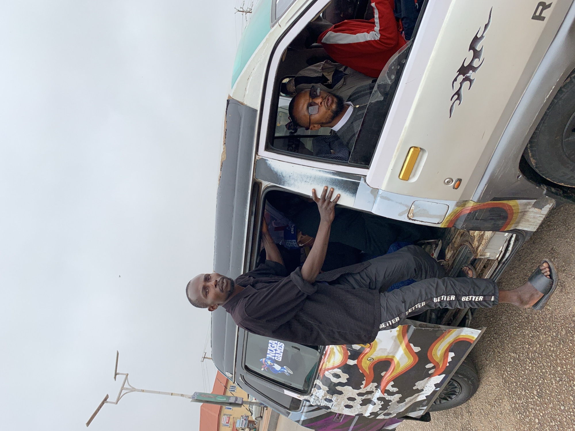 In the city of Gura Top, Nigeria, a man stands partially inside the open door of a passenger van, looking at the camera. He wears a black collared shirt and dark pants with white lettering that reads "BETTER BETTER BETTER." He has black sandals on. Another man, wearing sunglasses and a grey sweater over a white shirt, is in the driver's seat of the van. The van is white with colorful graffiti on its side. Other passengers are visible inside the van. The background shows a cloudy sky, a street lamp, and buildings.