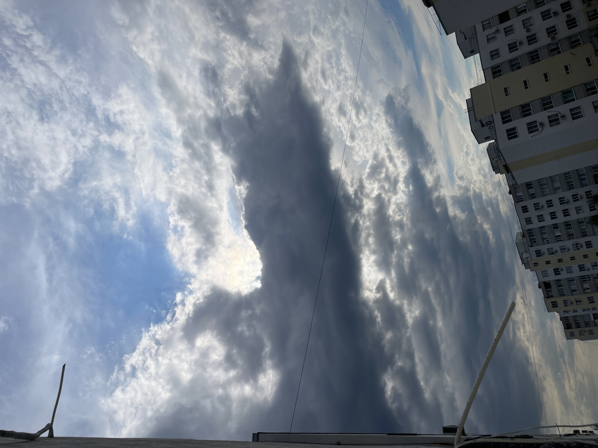 The image shows a cloudy sky above what appears to be a rooftop. The clouds are a mix of light and dark, with some patches of blue sky visible. A tall building is visible on the right side of the image. A pole is on the bottom right and bottom left, and a powerline crosses the center of the image. There is no evidence indicating the image was taken in Saratov, Russia. There are no people visible in the image.