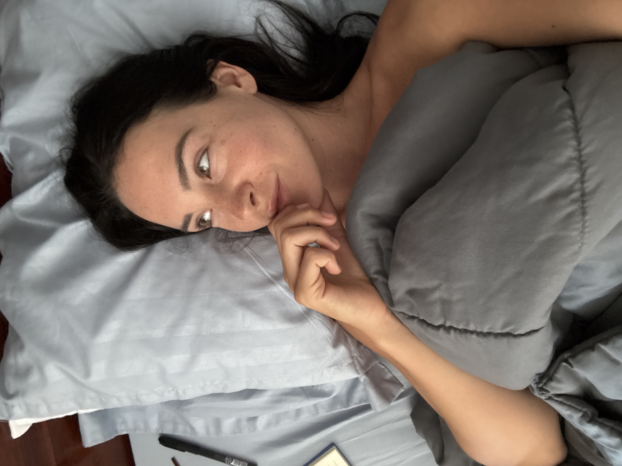 A woman with dark hair is lying in bed, propped up on a light blue, striped pillow. She is under a gray comforter, with her right arm resting on it. Her left hand is touching her chin as she looks to the right, her eyes open and her gaze directed upward. A black pen and a small object, possibly a book or notepad, are visible to her left on the bed. The scene appears to be a bedroom, with a wooden headboard or wall visible behind the pillow. The location is not specified in the image or its context.