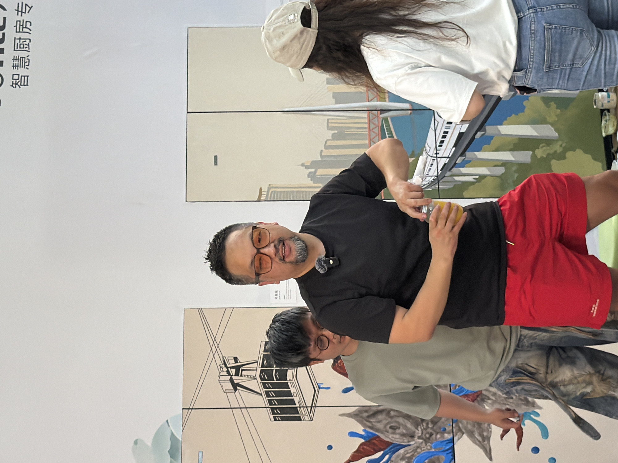 A man with short dark hair, a goatee, and orange-tinted glasses is holding a glass of orange liquid and appears to be pouring it into another container. He is wearing a black t-shirt and bright red shorts, with a small black microphone clipped to his shirt. He is standing in front of a large mural or display that features an artistic depiction of a city skyline with a bridge and a train. Behind him and to his left, another person with dark hair and glasses is partially visible, wearing a light green t-shirt and patterned pants. To the right of the main subject, a person wearing a light-colored baseball cap and a white t-shirt is facing away from the camera, with their back to the viewer. They are wearing blue jeans and have their hands at their sides. The background consists of a white wall with some Chinese text at the top and the large mural. The overall scene suggests an event or exhibition, possibly in a gallery or public space within Yuzhong District, China, given the context of the artwork.