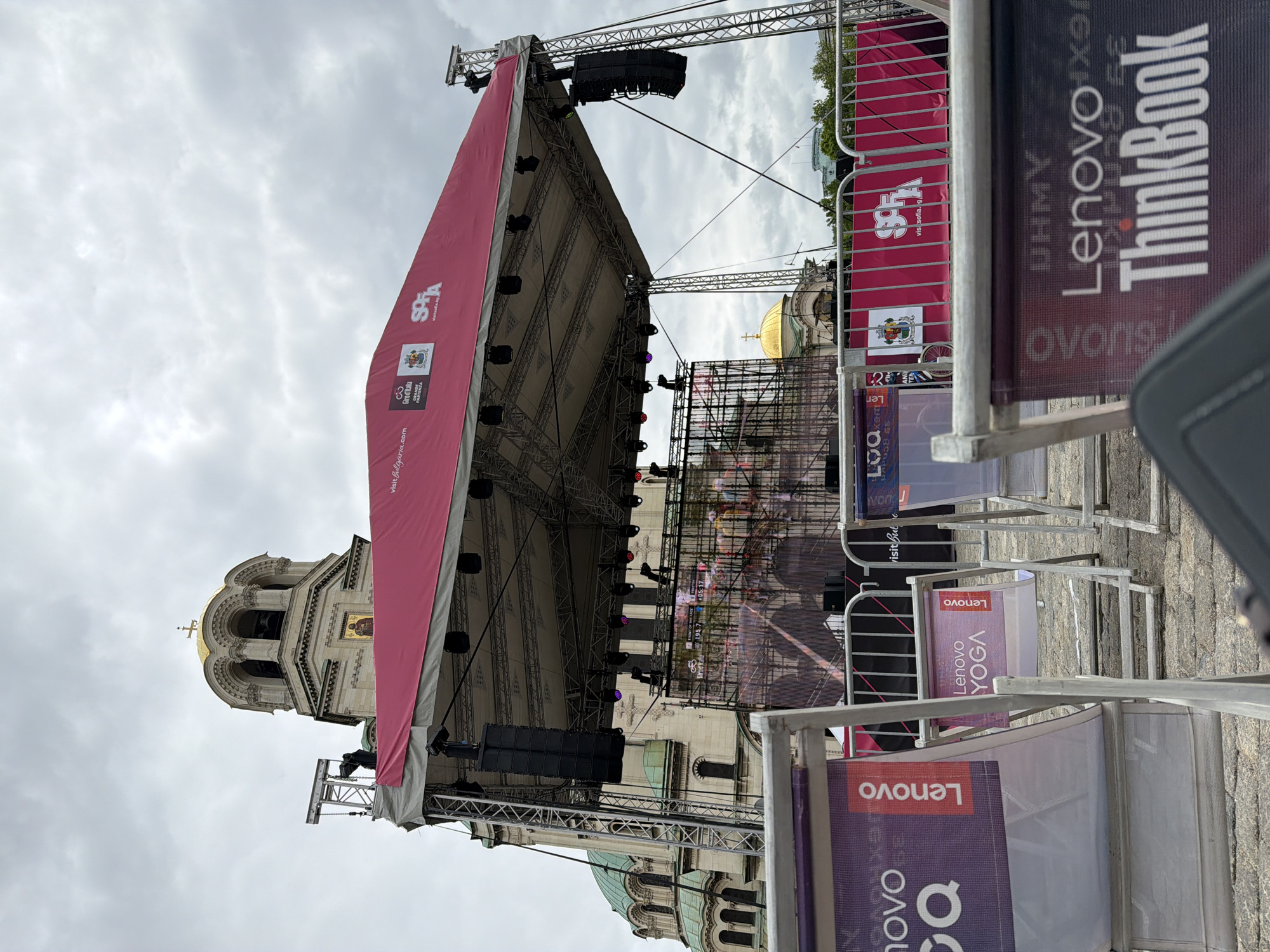 The image shows an outdoor event stage set up in front of the Alexander Nevsky Cathedral in Sofia, Bulgaria. The stage has a large red covering and a screen displaying images of cyclists. Several Lenovo advertisements are placed on barriers in front of the stage. The sky is overcast, and the cathedral's golden dome is visible in the background.