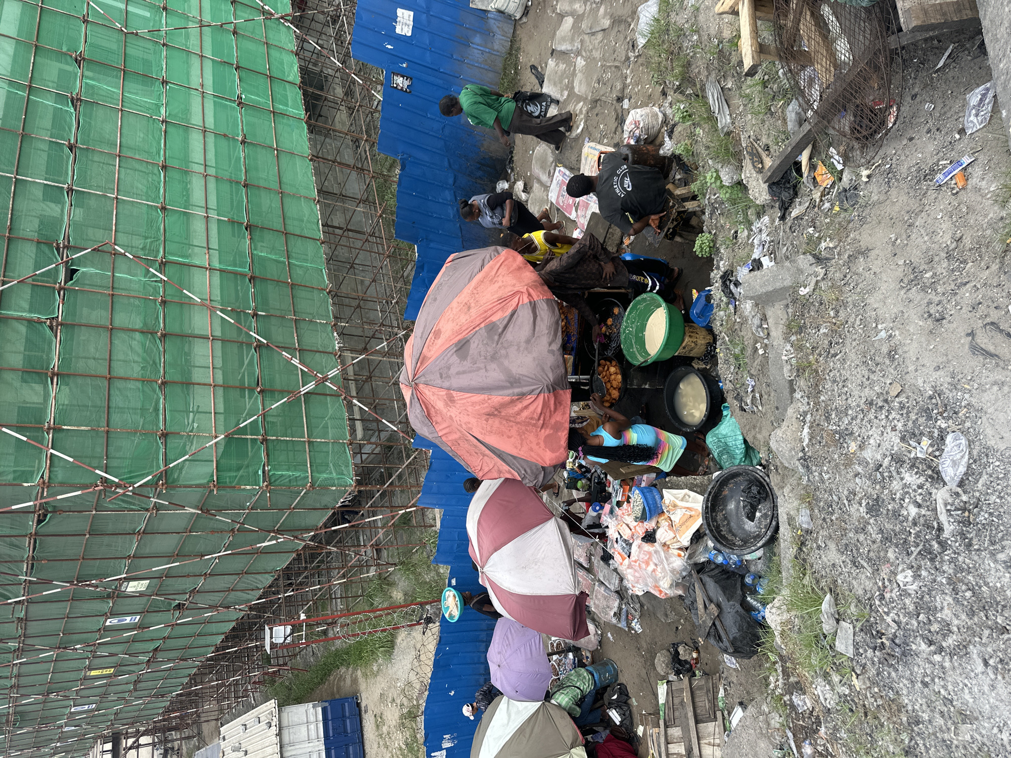 The image depicts an urban scene in Lagos, Nigeria. A large construction site with green mesh scaffolding dominates the left side of the image. Adjacent to the construction site is a bustling market area where several vendors are operating under colorful umbrellas. People are gathered around the vendors, suggesting commercial transactions are underway. The ground is cluttered with litter and debris. A blue corrugated metal fence borders the market area. The overall impression is one of a densely populated, informal commercial area adjacent to a construction zone in a developing urban environment.