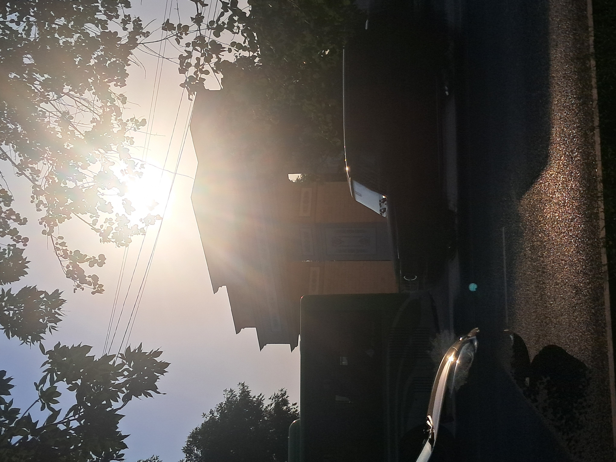 The image depicts a scene captured from a low angle, looking upwards and to the side. The sun is brightly visible, casting strong lens flares and illuminating the sky with a hazy glow, suggesting it is either sunrise or sunset. Silhouetted branches of leafy trees frame the upper left portion of the image. In the background, a building with a light brown facade and a distinct architectural roofline is visible. The building appears to be of traditional design, possibly a house or a public building.
In the foreground and right side of the image, the scene is dominated by dark, indistinct objects. Part of a car is visible, with a shiny chrome detail catching the sunlight. A darker, larger object, possibly another vehicle or a piece of equipment, is also present. The ground is a textured asphalt surface, illuminated by the sunlight on the right side, creating a dappled effect. The overall mood is serene, with the intense sunlight creating a dramatic and atmospheric quality.
There is no visible text in the image. The location context provided, Shymkent, Kazakhstan, does not offer specific visual cues that are definitively identifiable within the image itself. The time of day is suggested by the low sun angle and the quality of light.