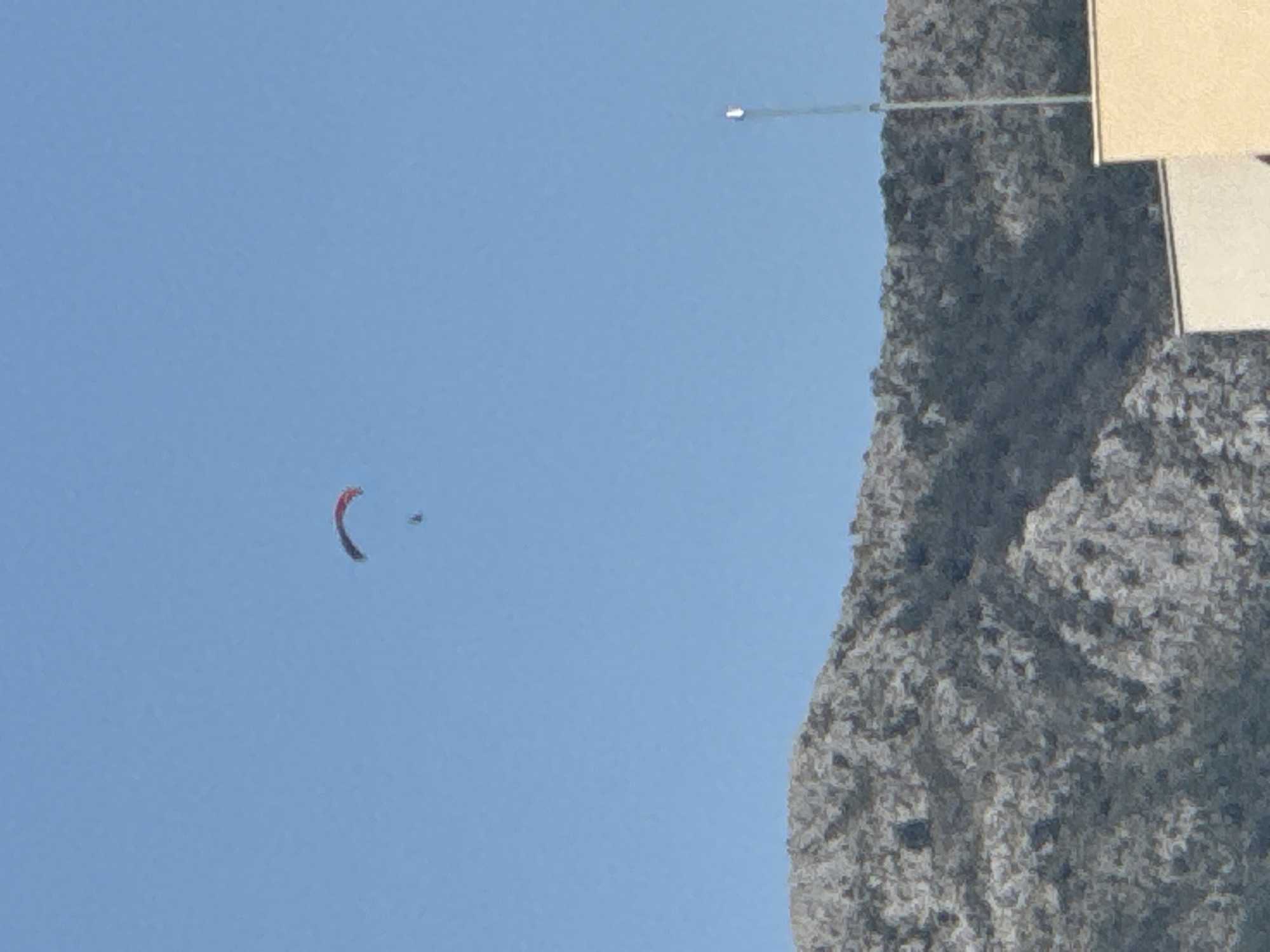 A lone paraglider is visible in the clear blue sky above a rocky mountain range. The paraglider, with red and black elements, is suspended in the air, with a small figure attached. The mountain range, which appears rugged and sparsely vegetated, forms the lower portion of the image. In the foreground, the top of a light-colored building is visible, with a thin pole extending upwards into the sky on the right side of the frame. This scene likely depicts an activity occurring near Budva, Montenegro, given the characteristic mountainous terrain and coastal proximity suggested by the clear sky.