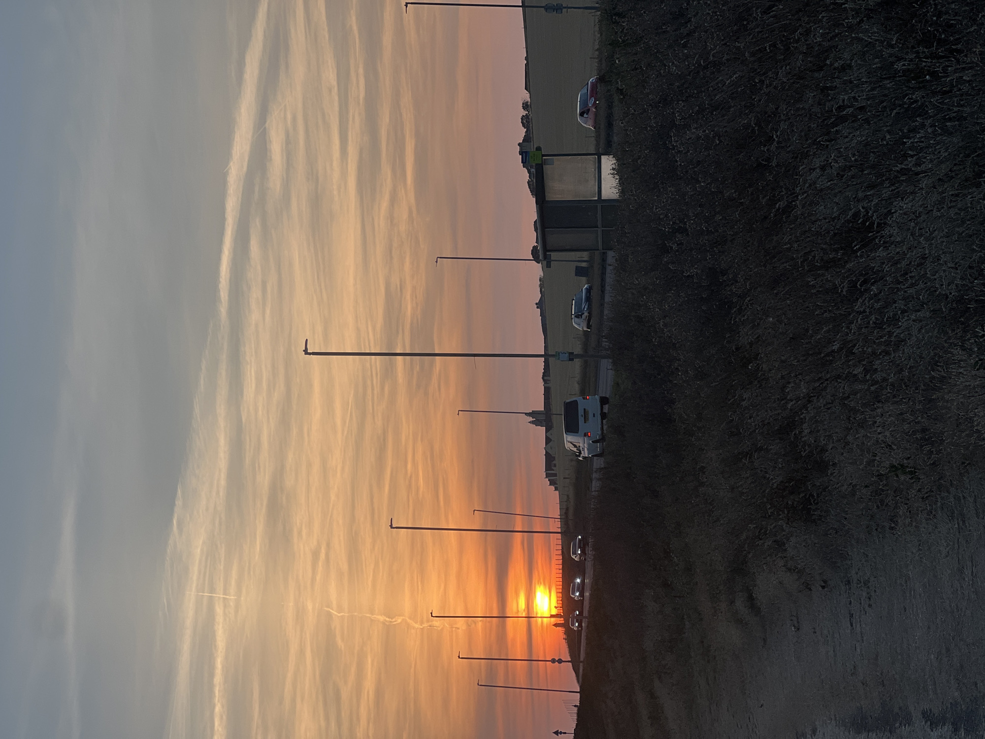 The image depicts a road scene at sunset in Ovingdean, United Kingdom. A bright orange sun is low on the horizon, casting a warm glow across the sky, which transitions from orange to pink and then to a pale blue. Wispy clouds, some with contrails, are scattered across the sky.
Several cars are visible on the road, including a white van on the right side, and three cars in the distance driving towards the sunset. There are multiple tall lampposts lining the road. On the right side of the road, a bus shelter with glass panels stands beside the road. In the background, rolling hills and buildings can be seen, suggesting a rural or semi-rural setting. The foreground is dominated by dark, silhouetted vegetation, possibly bushes and long grass, along a dirt path on the left. The overall impression is a peaceful, end-of-day scene on a road leading towards a picturesque landscape.