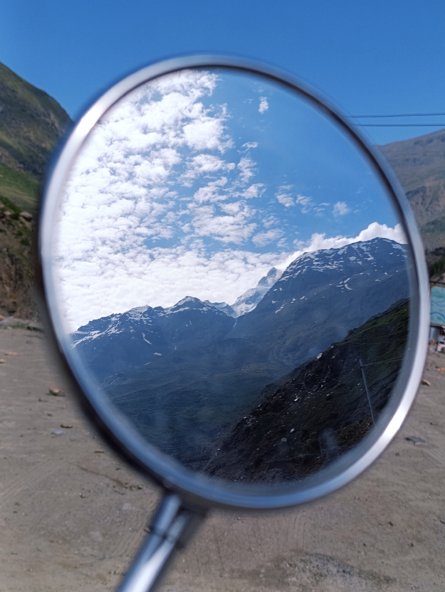 The image shows a mirror reflecting a mountainous landscape under a blue sky with scattered clouds. The reflection captures snow-capped peaks and green slopes. In the foreground, outside the mirror, is a gravelly surface and the metallic handle of the mirror, suggesting it is a rearview mirror of a vehicle. The sky is bright and clear, indicating daytime. The location is identified as Sissu, India, a region known for its scenic Himalayan vistas.