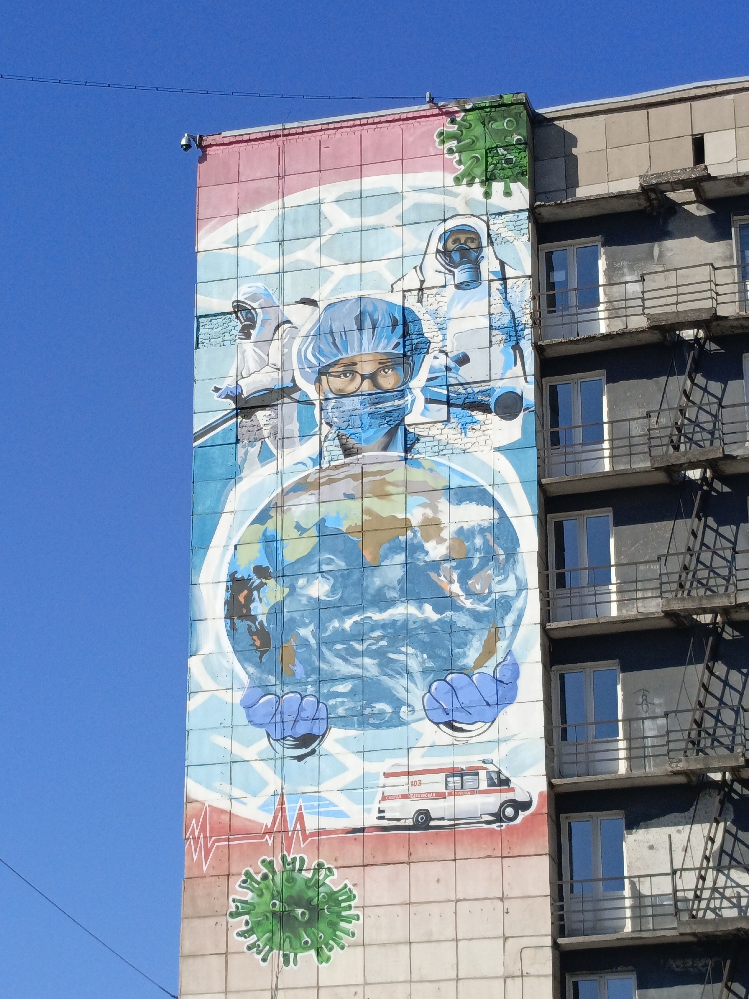 This image depicts a mural on the side of a building in the city of Perm, Russia. The mural is painted on large, square tiles and features medical personnel in protective gear. A central figure wears a surgical cap, mask, and glasses, holding a globe. Two other figures in hazmat suits are positioned behind the central figure. At the bottom of the mural, there is a drawing of a green virus, an EKG heartbeat line, and an ambulance. A similar virus drawing is in the upper right corner. The building adjoins the mural, with visible windows, balconies, and fire escapes. The sky is blue. A security camera is present at the top of the mural.