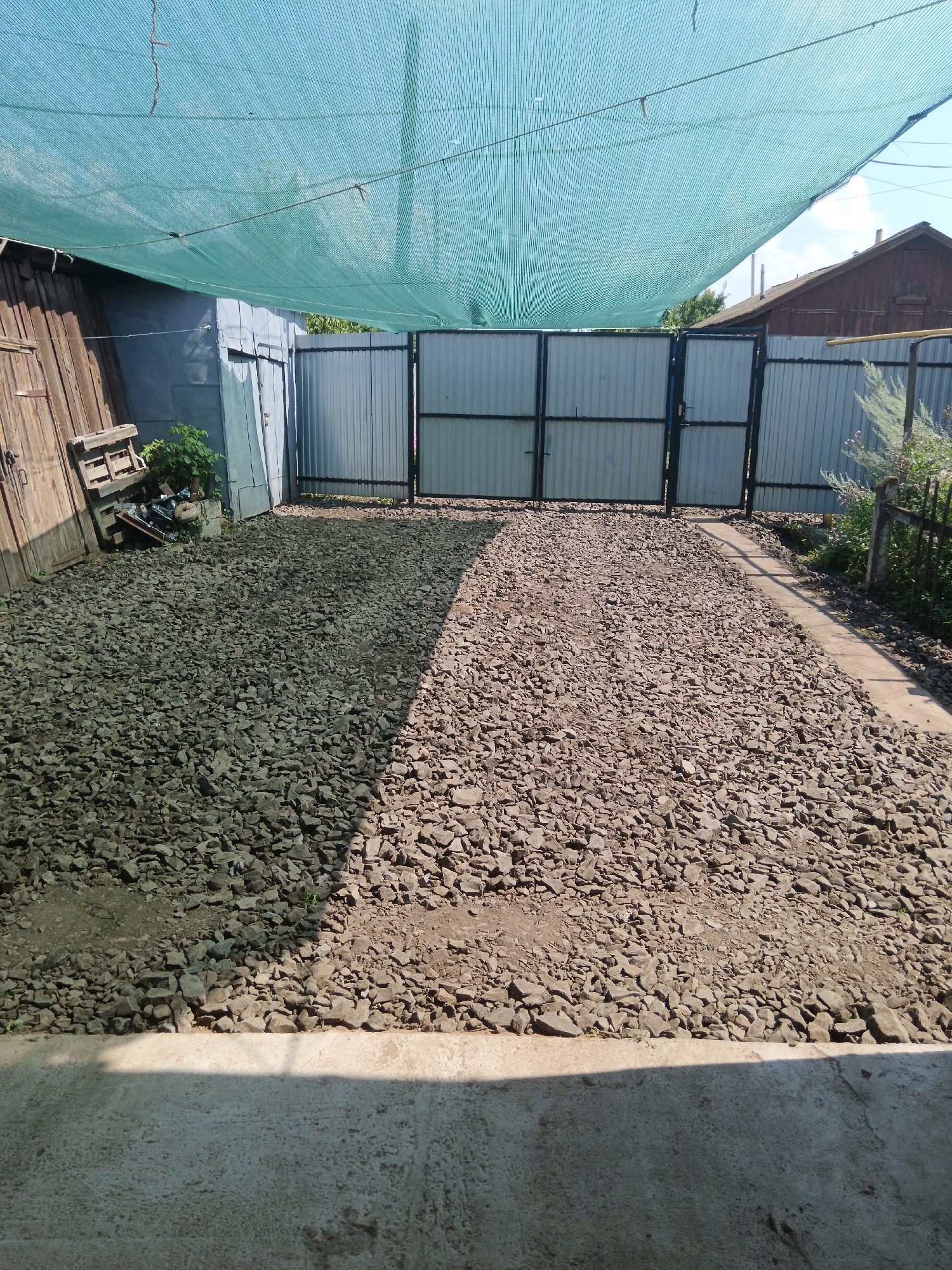 This image depicts an outdoor scene, possibly a backyard or a service area, in Sorochinsk, Russia. The foreground features a concrete surface with a shadow cast across it from an unseen structure. Beyond the concrete, the ground is covered in a thick layer of gravel and small stones, forming a pathway or a yard.
In the middle ground, a dark metal gate with vertical panels and a horizontal crossbar stands centrally. To the left of the gate, there is a wooden structure with a door and a wooden pallet stacked with what appears to be debris or materials. A small green plant grows near the wooden structure. To the right of the gate, a fence made of corrugated metal panels extends outwards, and beyond that, a section of a grey metal fence with vertical bars is visible. Further to the right, a dark metal fence with ornate detailing and a concrete post can be seen, with some dry vegetation in front of it.
The background shows the roofline of a dark wooden building with a visible chimney, and a glimpse of the sky with clouds. A large, light blue or green mesh tarp is stretched across the top of the image, supported by cables, suggesting it's used for shade or protection. The overall impression is one of a functional outdoor space, possibly undergoing some form of renovation or maintenance.