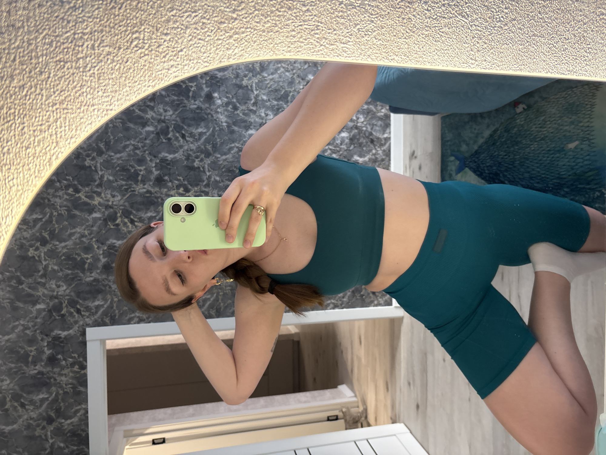 A young woman is captured in a mirror selfie, posing indoors in Izhevsk, Russia. The image is rotated 90 degrees clockwise, depicting her as if lying on her left side, holding a light green smartphone with two visible camera lenses to take the picture. She wears a matching teal or dark green athletic set, consisting of a sports bra and high-waisted cycling shorts with a subtle ribbed texture. A small, dark rectangular tag is visible on the left hip of her shorts. She has brown hair styled in a low braid or ponytail over her shoulder, and wears a golden ring with an intricate design on her right ring finger, along with a delicate gold necklace featuring a small heart-shaped pendant. A white sock covers her right foot.
The mirror she is using has a distinctive, illuminated frame with a rough, textured surface along the top and left edges, casting a warm glow. Reflected in the mirror behind her is a dark grey wall with a white marble-like pattern. Further back in the reflection, a bed with a light blue duvet or blanket is visible, along with what appears to be a blue rug or floor covering featuring a decorative, scale-like pattern, possibly resembling a mermaid tail. Parts of white doorframes or furniture are also reflected, as is a wooden-textured wall or surface to the right. Her facial expression is neutral as she looks towards the phone screen.