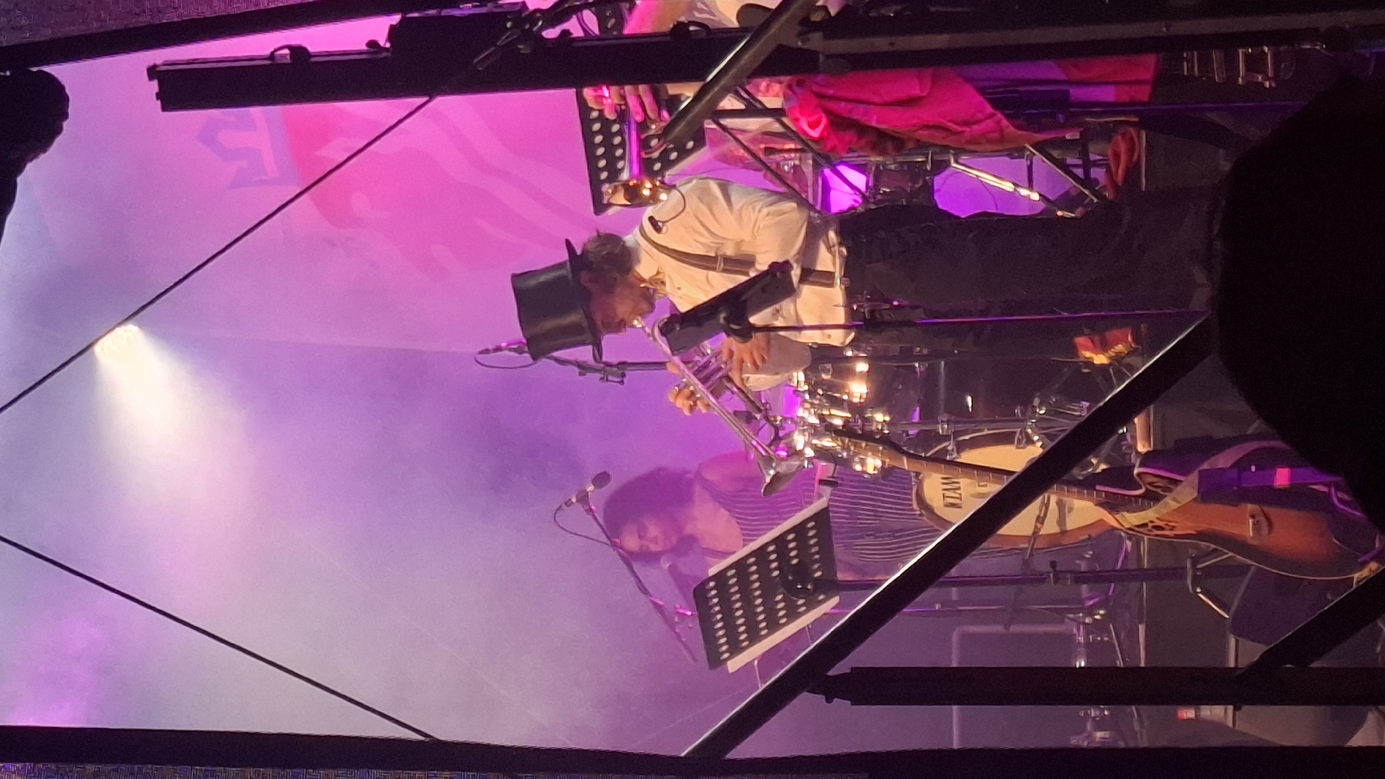 A band performs on a dimly lit stage in Differdange, Luxembourg. A man in a top hat plays a trumpet, wearing a white shirt, suspenders, and dark pants. To his left, a woman with dark curly hair sings into a microphone, wearing a striped dress. In front of them are drums and a guitar, with sheet music stands positioned strategically. The stage is illuminated by purple and pink lights, with smoke creating a hazy atmosphere. The background features a large banner with a crown motif. The perspective is from the audience, with parts of the stage structure in the foreground.