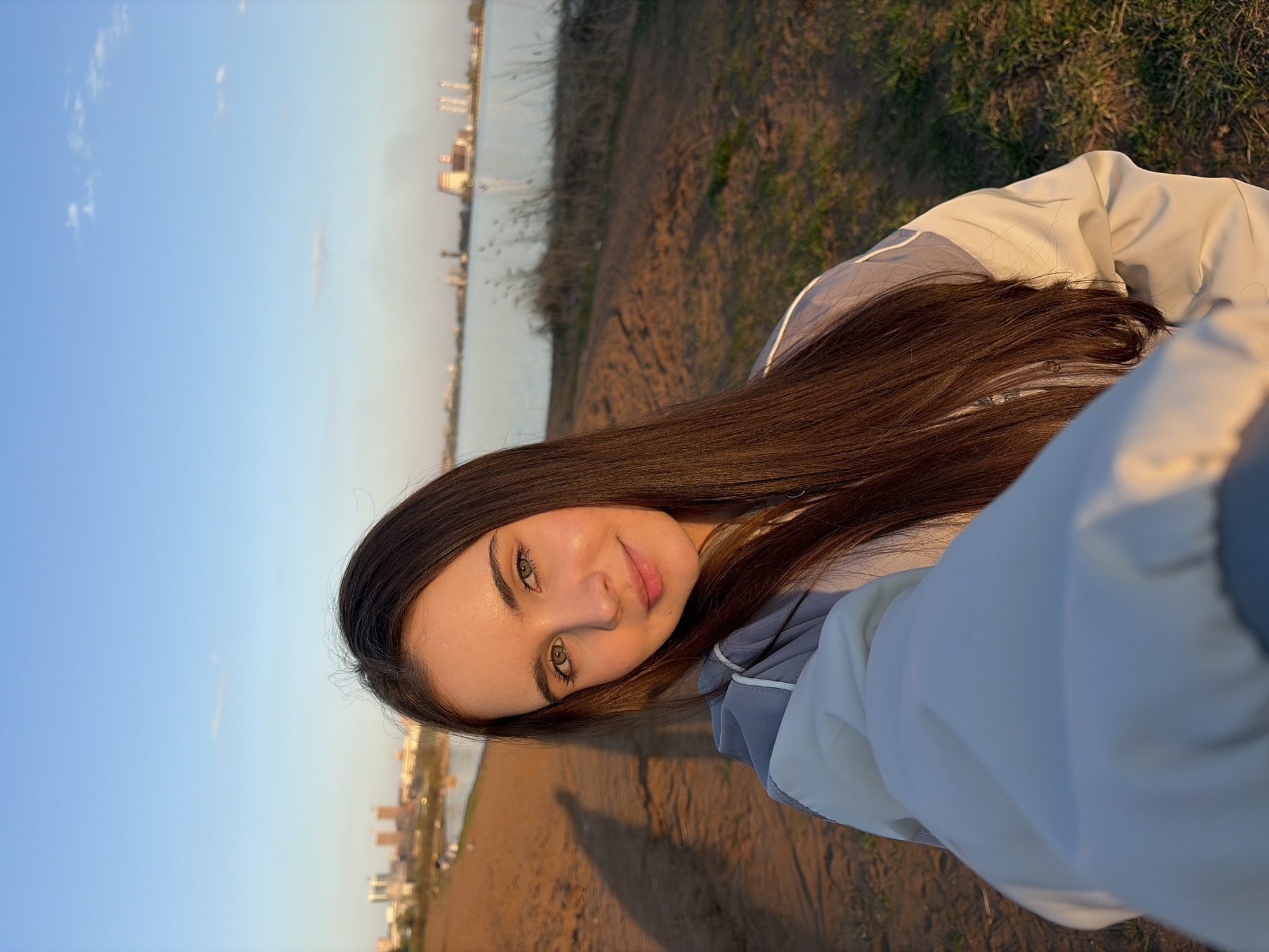 A young woman with long, dark brown hair is posing for a selfie outdoors. Her gaze is directed toward the camera. She is wearing a light-colored jacket with gray accents. The background consists of a riverbank, water, and a distant cityscape that may be Izhevsk, Russia. The sky is blue with a few clouds, suggesting it may be sunset. A long shadow is visible on the riverbank near the bottom of the image.
