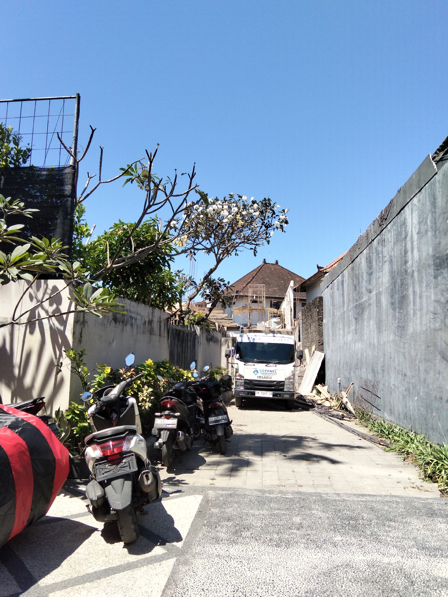 The image shows a narrow paved alleyway in Denpasar, Indonesia, on a clear, sunny day. A white Isuzu truck with the logo "DEPANEL" and Indonesian license plate "DK 3081 CA" is parked in the middle of the alley. The truck appears to be part of a construction or delivery service.
To the left of the truck, a row of parked motorcycles is visible. A large, red and black striped inflatable object is positioned in the foreground on the left. Behind the motorcycles and truck, there is a concrete wall on the right side of the alley, and on the left, a wall with plants and trees growing over it. A traditional Indonesian-style house with a tiled roof is visible further down the alley in the background.
The alleyway is lined with buildings and vegetation, suggesting it is a residential or commercial area. The overall scene is one of an ordinary day in Denpasar, with elements of both daily life (motorcycles) and economic activity (the truck and construction materials).
