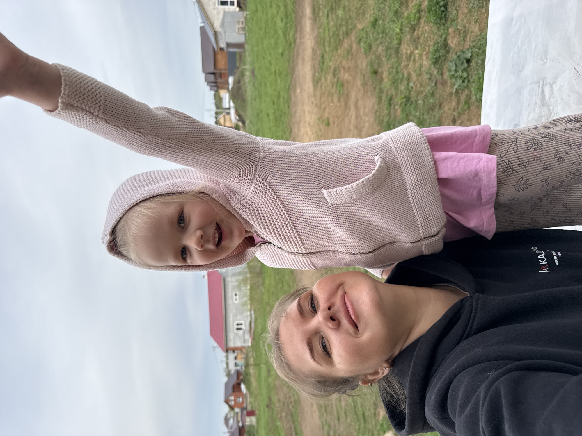 A young woman and a small child are in a selfie outdoors. The woman is in the foreground, wearing a black hooded sweatshirt with the words "HR KADRO RECRUITI PERM" on the right chest, looking up at the camera with a slight smile. Above her and to the left, a small child with blonde hair wearing a pink hooded sweatshirt with a kangaroo pocket smiles and extends one arm upwards. The background shows a rural area with green grass, a dirt road, and several buildings, including a white building with a red roof and wooden houses with brown roofs. The image is taken from a low angle. This image was taken in Ферма, Russia.