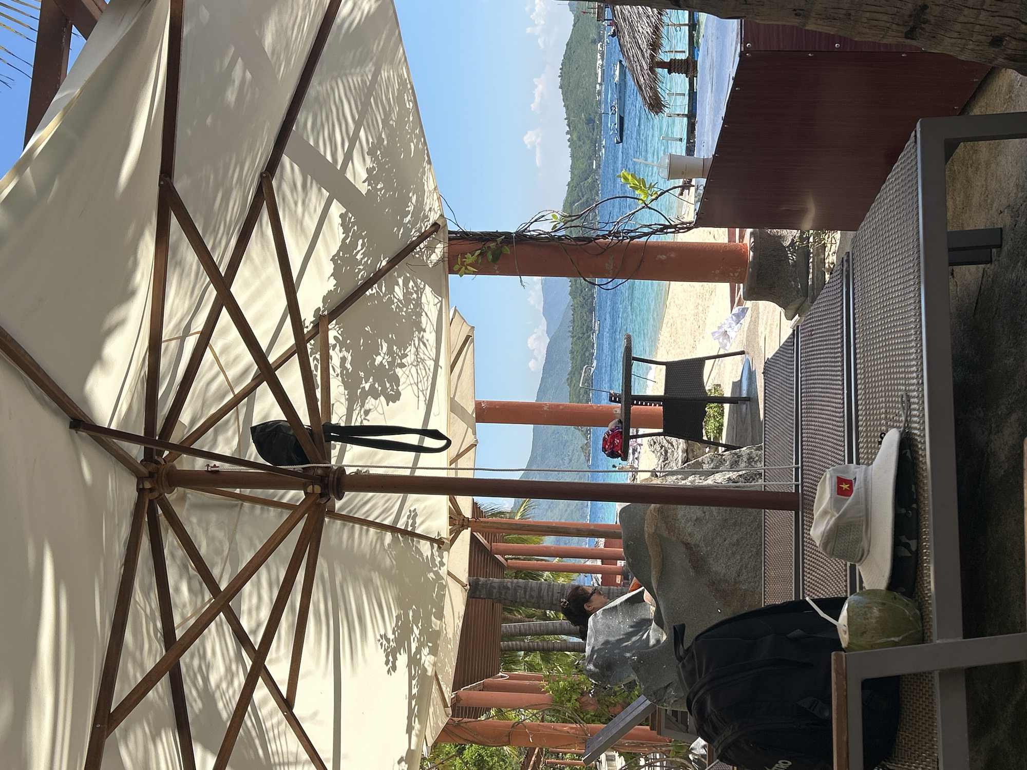 Under a large white umbrella, a beach scene unfolds. In the foreground, a black backpack rests on a lounge chair next to a halved coconut with a straw. A white hat with a Vietnamese flag emblem sits on top of a folded black fabric. In the middle ground, a person sits on a rocky formation, looking towards the sea. Behind them, a beach with fine sand meets the turquoise ocean, dotted with several boats. Mountains form a distant backdrop under a clear blue sky with scattered clouds. The scene is located at a beachside resort in Nha Trang, Vietnam, indicated by the hat's emblem.