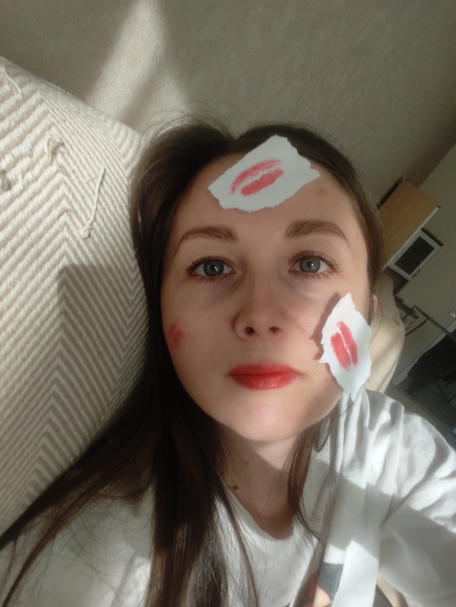 A woman with fair skin and brown hair is pictured indoors. She is wearing red lipstick and has three pieces of torn white paper stuck to her face, each bearing a red lipstick mark in the shape of lips. One paper is on her forehead, another on her right cheek, and the third near her left cheek. She appears to be lying down, leaning against a beige and white patterned cushion or headboard. The background includes a white ceiling and a glimpse of a cabinet or appliance. The location is Unknown, Unknown.