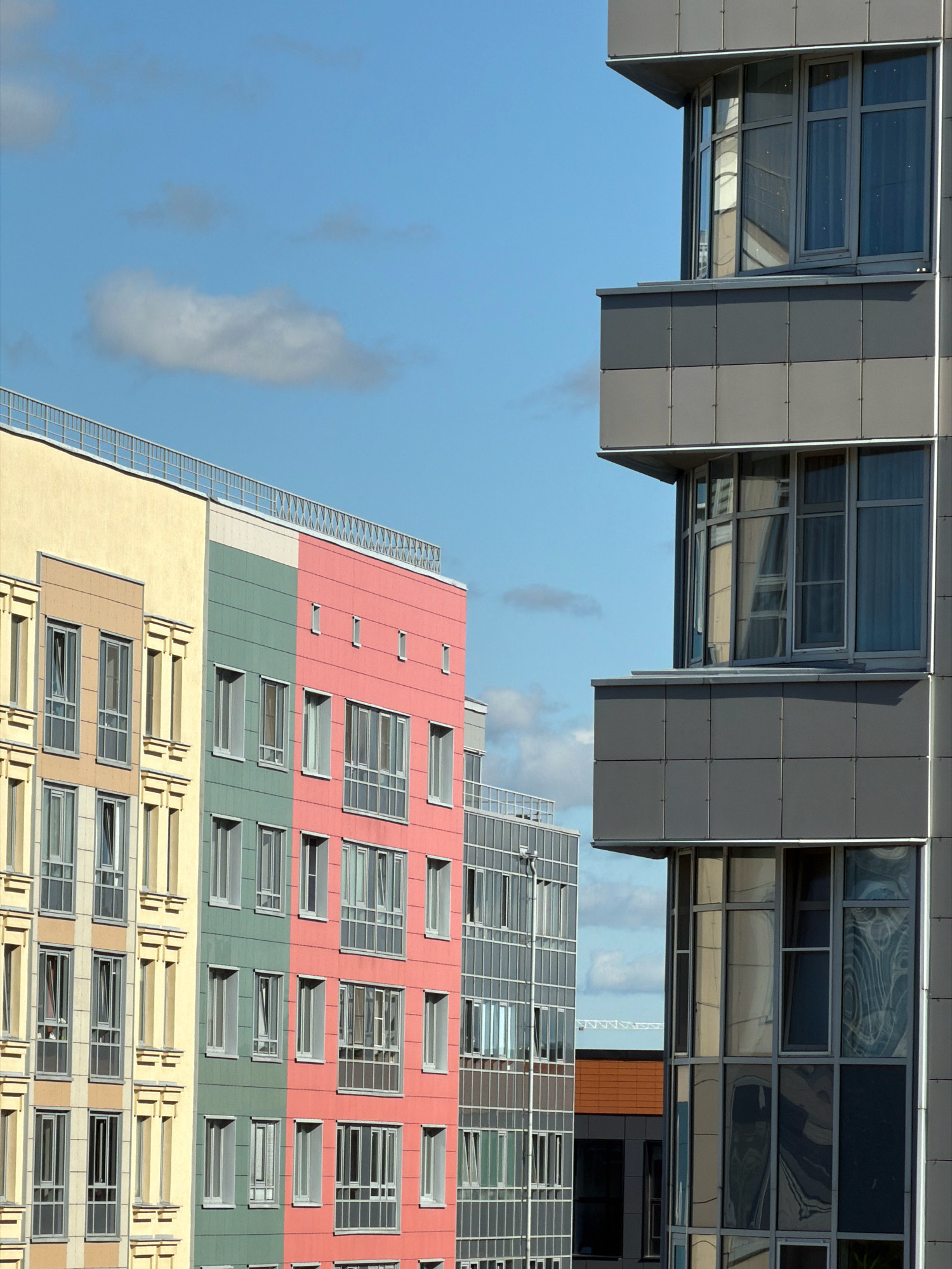 This is a medium shot of a modern residential building facade against a partly cloudy blue sky. The building is segmented into different color blocks. On the left, a section of the building is a pale yellow, followed by a section in a light brown color with decorative elements. Next to it, a section of the building is colored a vibrant green, and further to the right is a section in bright pink. These colored sections have multiple windows. To the far right of the image, a different section of the building, possibly a different structure, is clad in gray panels with large windows, some of which reflect the sky and adjacent buildings. The windows are multi-paned, with some appearing to be open. The top of the buildings features railings or decorative elements. The scene is devoid of people and action, presenting a static architectural view. The presence of the building and its modern design is consistent with new construction found in areas like the Primorsky District of Saint Petersburg, Russia, known for its contemporary residential complexes.