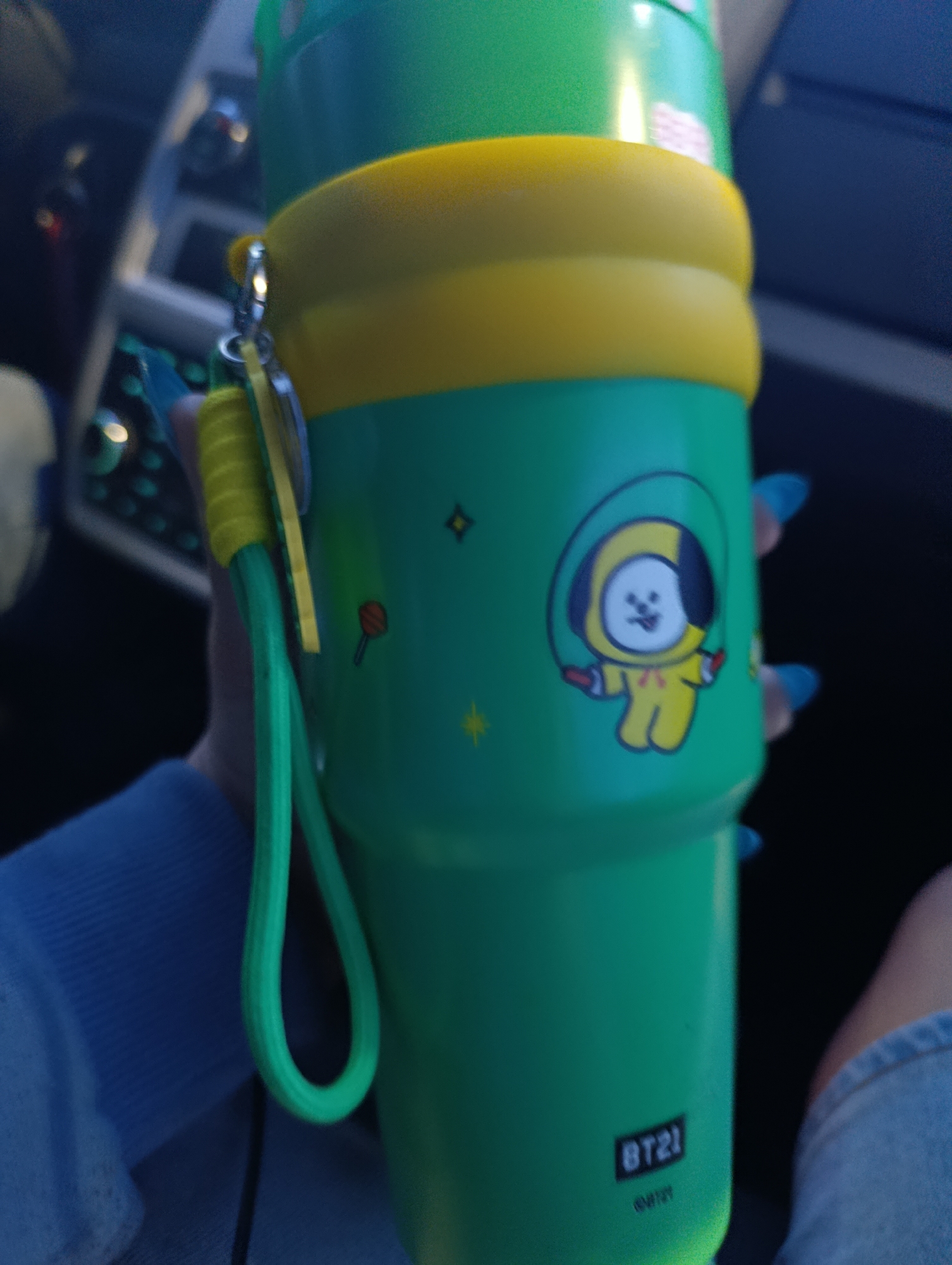 A close-up, eye-level shot captures a person's hand holding a vibrant green and yellow tumbler inside a vehicle in Funza, Colombia. The person wears a blue long-sleeved top, and their fingers are adorned with bright blue nail polish.
The primary subject is a tall, green tumbler featuring a wide yellow band around its center. A green wrist strap, attached to a yellow clip, dangles from the top section of the tumbler. On the front, there is a clear illustration of Chimmy, the yellow character from BT21, depicted in a hooded tracksuit, sticking its tongue out, and holding a jump rope. Around the character are small graphics including a red lollipop, yellow stars, and a black diamond. Below the illustration, the "BT21" logo is prominently displayed, with the word "CHIMMY" printed beneath it in smaller text.
The background is softly blurred but reveals elements of a car's interior, including parts of the dashboard and controls, suggesting the person is either traveling or parked. The lighting is diffused, indicating daytime conditions inside the vehicle. The image focuses on the personalized tumbler and the hand holding it, presenting a casual moment.