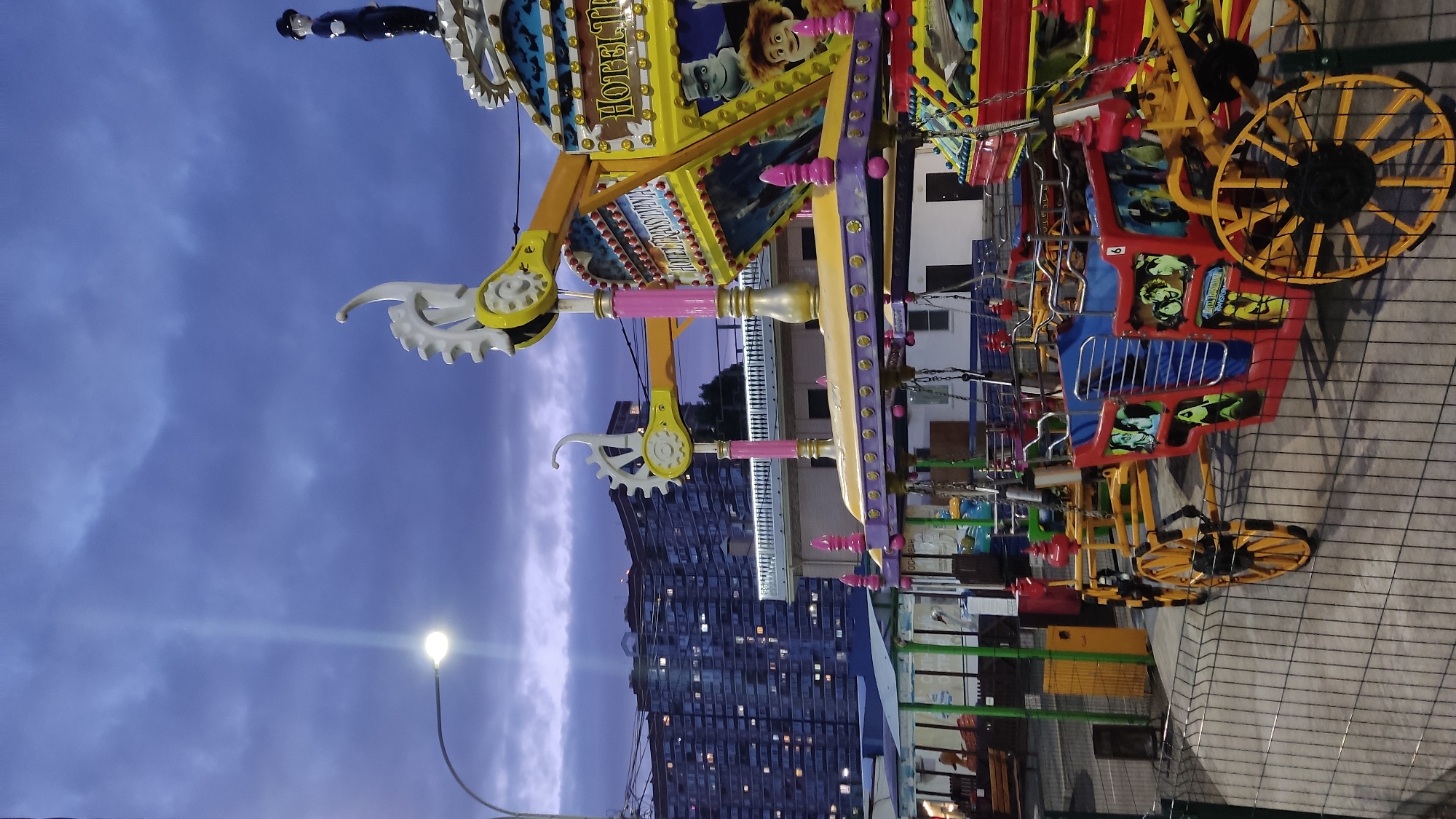 The image depicts a colorful amusement park ride at dusk, possibly in the city of Krasnodar, Russia. The ride, themed around "Hotel Transylvania," features several carriages decorated with characters from the movie. A tall apartment building with many illuminated windows is visible in the background, suggesting an urban setting. A street lamp on the left illuminates the scene with a bright light against the dark, cloudy sky. The ride appears to be stationary, with no people actively riding it. The overall atmosphere is one of an evening at an amusement park as night begins to fall.