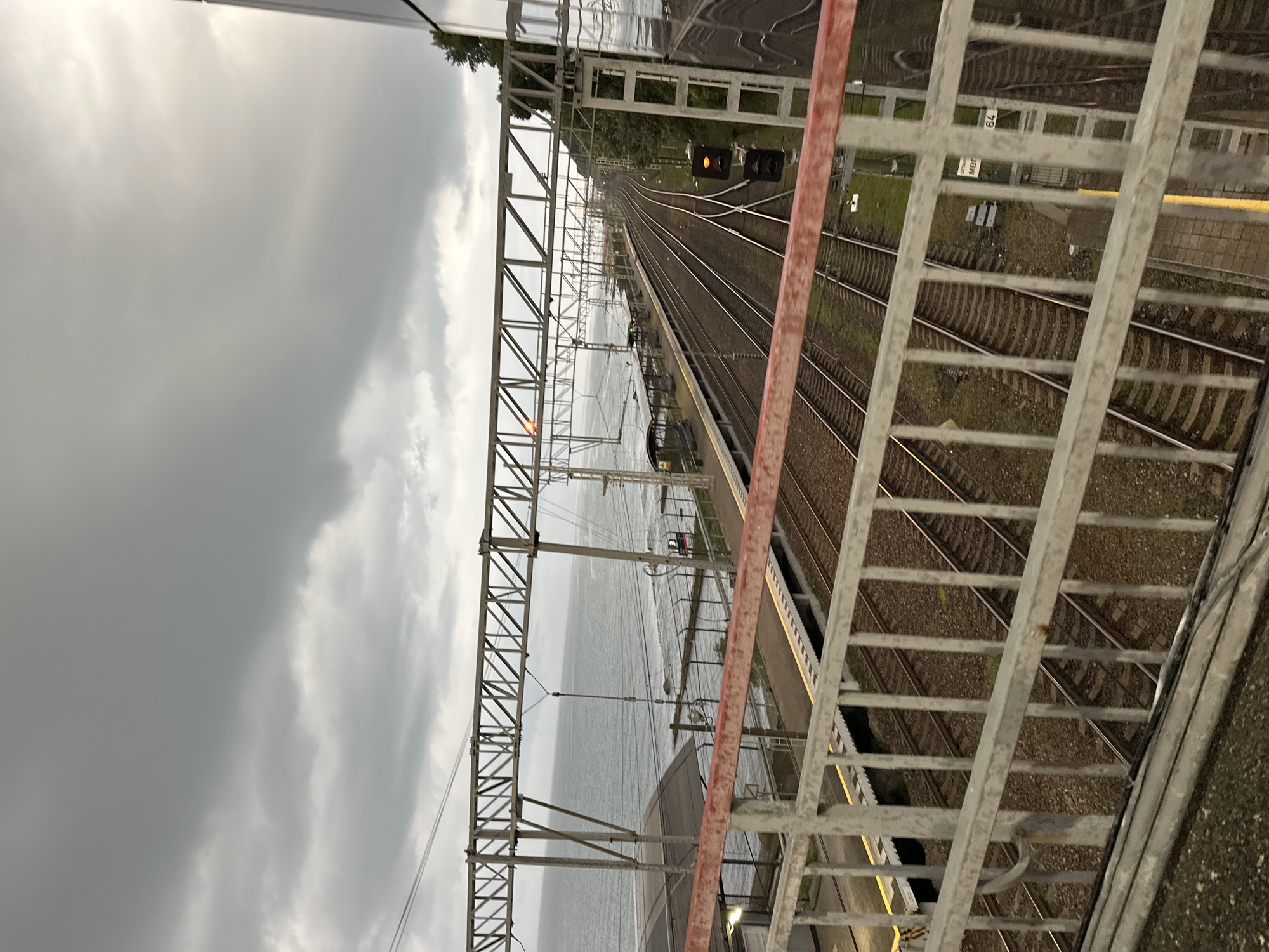 The image shows a view of a train station and railway tracks in Sochi, Russia. The perspective is from an elevated walkway, looking down at multiple parallel train tracks that curve into the distance. To the left of the tracks, there is a body of water, likely the Black Sea, with a shoreline and a fence. Overhead, there are numerous metal support structures for power lines and other infrastructure. The sky is overcast with heavy, gray clouds. A red railing runs across the foreground of the image. There are no people visible in the scene. The train station platform is visible with covered shelters and yellow safety markings along the edge of the platform. A traffic signal with an orange light is visible on a pole next to the tracks.