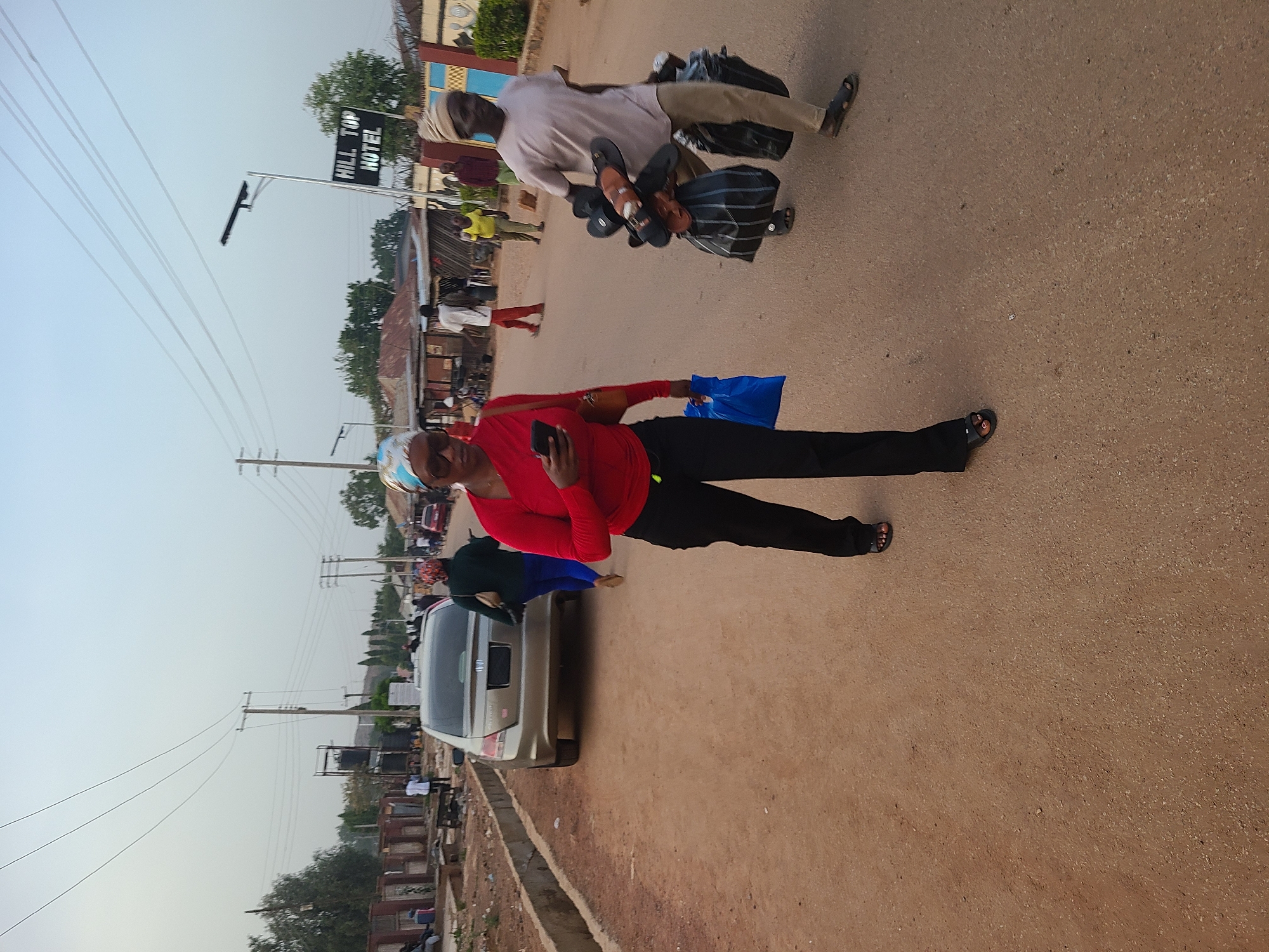 The scene is on a street in Jos, Nigeria. A woman in a red long-sleeved shirt and black pants stands holding a phone and a blue plastic bag. Nearby, a man in a light-colored shirt and pants carries bags and footwear. A silver sedan is parked beside the road. In the background, there are utility poles, overhead wires, buildings, and people. A sign for "Hill Top Hotel" is visible. The road surface appears paved.