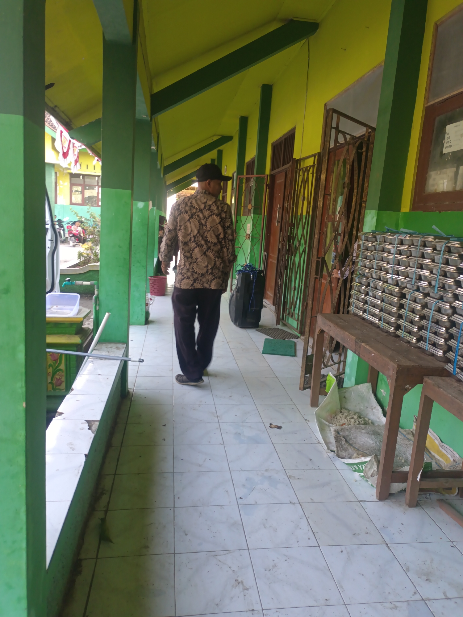 The image depicts a man walking down a covered walkway, possibly in a school or community center in Sarimulyo, Indonesia. The walkway has a green painted ceiling and pillars, with yellow walls visible. The floor is tiled with white and light grey squares. To the left of the walkway, there is a low green railing with decorative floral motifs painted on it. A white plastic container with a blue lid is placed on the railing. Further down the walkway, a dark blue speaker is leaning against the wall. To the right, a stack of metal trays, neatly tied with blue straps, is placed on the ground. A rustic wooden bench is situated in front of the stack of trays, with a large white bag filled with white material placed underneath it. In the background, metal gates and doors are visible, suggesting entrances to rooms. Several motorcycles are parked on the left side of the walkway, and a few people can be seen in the distance. The overall impression is of an outdoor communal space.