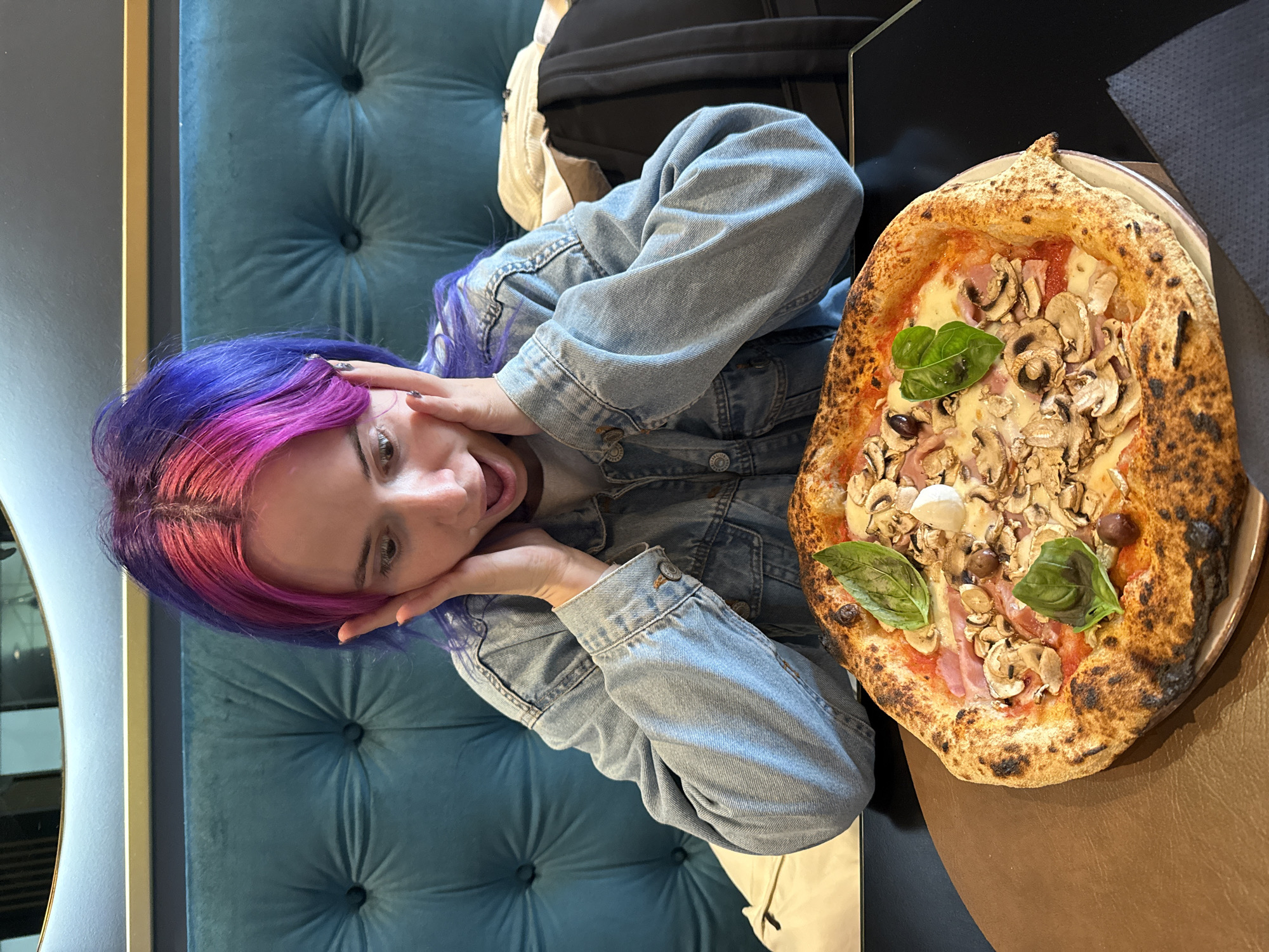 A young woman with brightly dyed purple and pink hair sits at a table with a large pizza in the City of Zagreb, Croatia. She wears a denim jacket and holds her hands to her face with a surprised expression. The pizza has a charred crust and is topped with mushrooms, basil, and other ingredients. The table is dark, and a tufted blue chair is visible behind the woman. A black bag sits nearby.