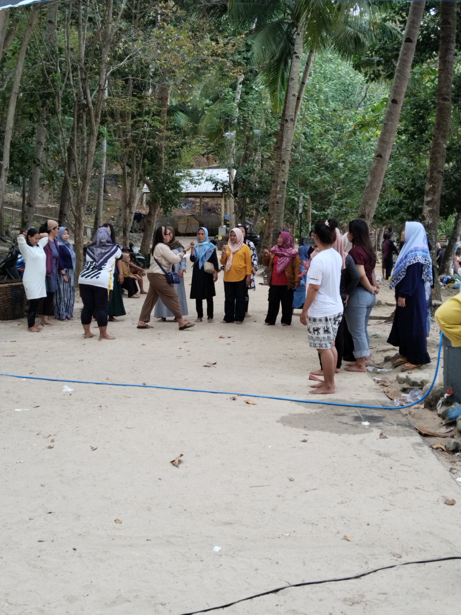 A group of people, most of whom are women wearing Islamic headscarves, stand on a sandy surface near a wooded area with palm trees. The group appears to be gathered for an event or activity. Some individuals are talking to each other, while others are looking in different directions. The environment suggests a tropical or subtropical location, possibly a beach or a coastal area in Prigi, Indonesia, given the presence of sand, palm trees, and a rustic, outdoor setting. There is a blue hose lying on the ground, indicating potential water access or a setup for some activity. In the background, a simple structure with a metal roof can be seen, suggesting some form of shelter or building. The overall scene conveys a casual, communal gathering.