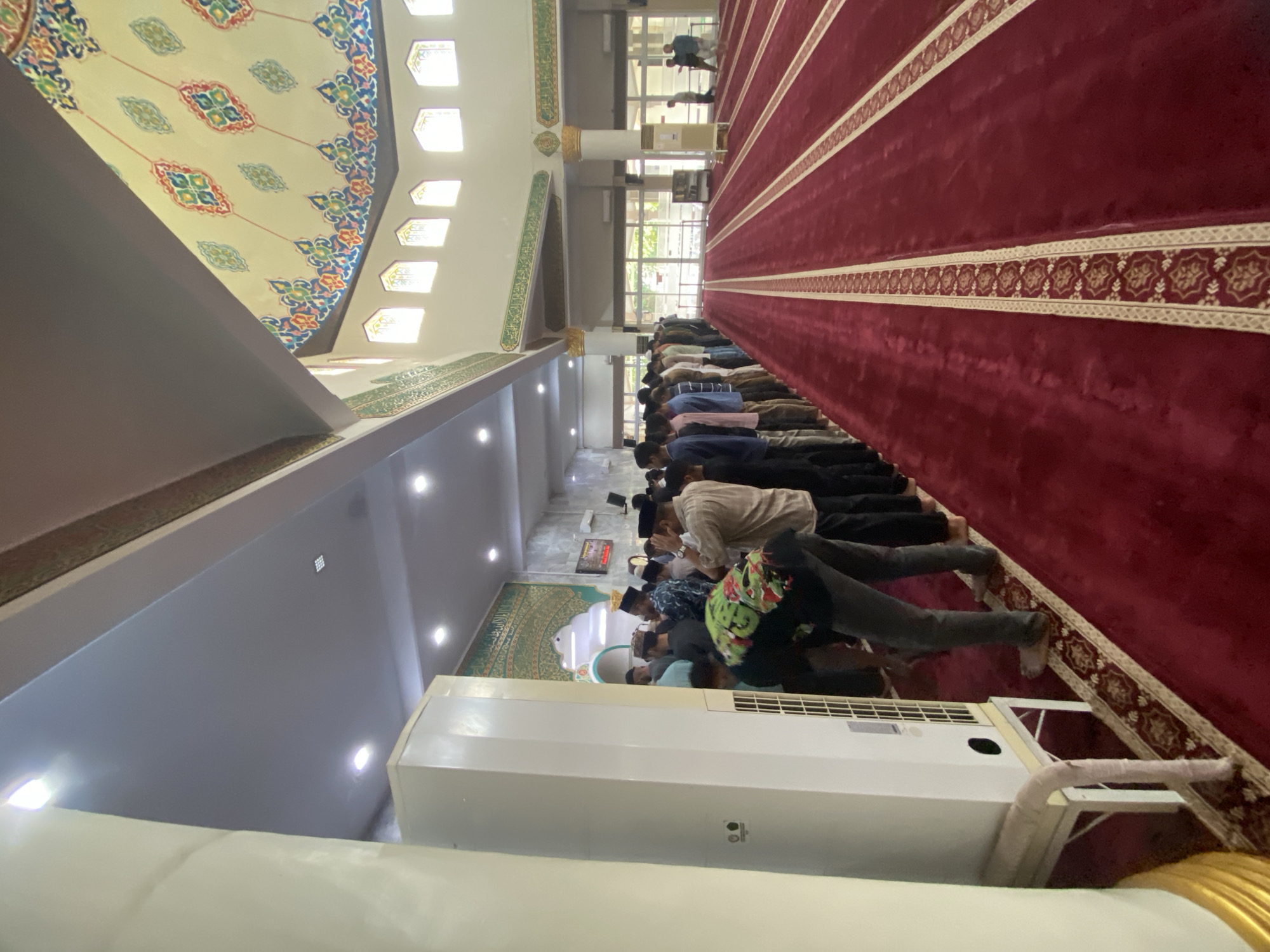 The image shows the interior of a mosque, likely in Lhokseumawe, Indonesia, during a prayer service. A long line of men, dressed in modest attire and some wearing skullcaps, are standing in prayer formation on a red carpet. The carpet has a patterned border. To the left of the image, a tall, white air conditioning unit stands beside a large, cream-colored pillar. The ceiling features intricate Islamic geometric patterns in vibrant colors and Arabic calligraphy. Natural light enters through arched windows along the upper wall. Another group of men can be seen in the background, some standing and others in prayer. The overall impression is one of a community gathered for religious observance in a well-maintained and decorated place of worship.