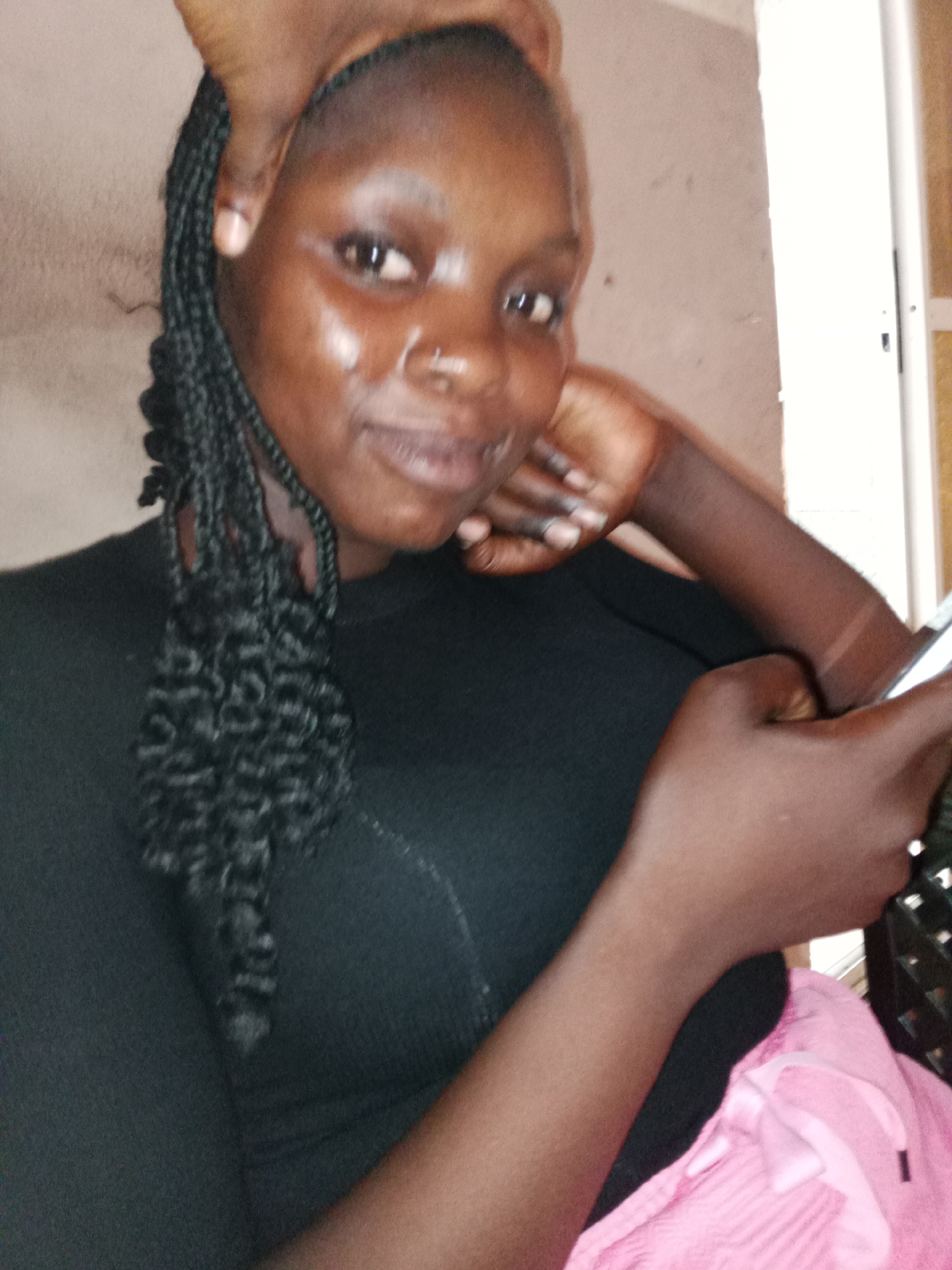 A young Black woman with braided hair, wearing a black shirt and pink bottoms, is holding a phone in her right hand and leaning her head on her left hand. Her right hand is on her cheek. The background is a plain wall. The image is likely taken in Jalingo, Nigeria.