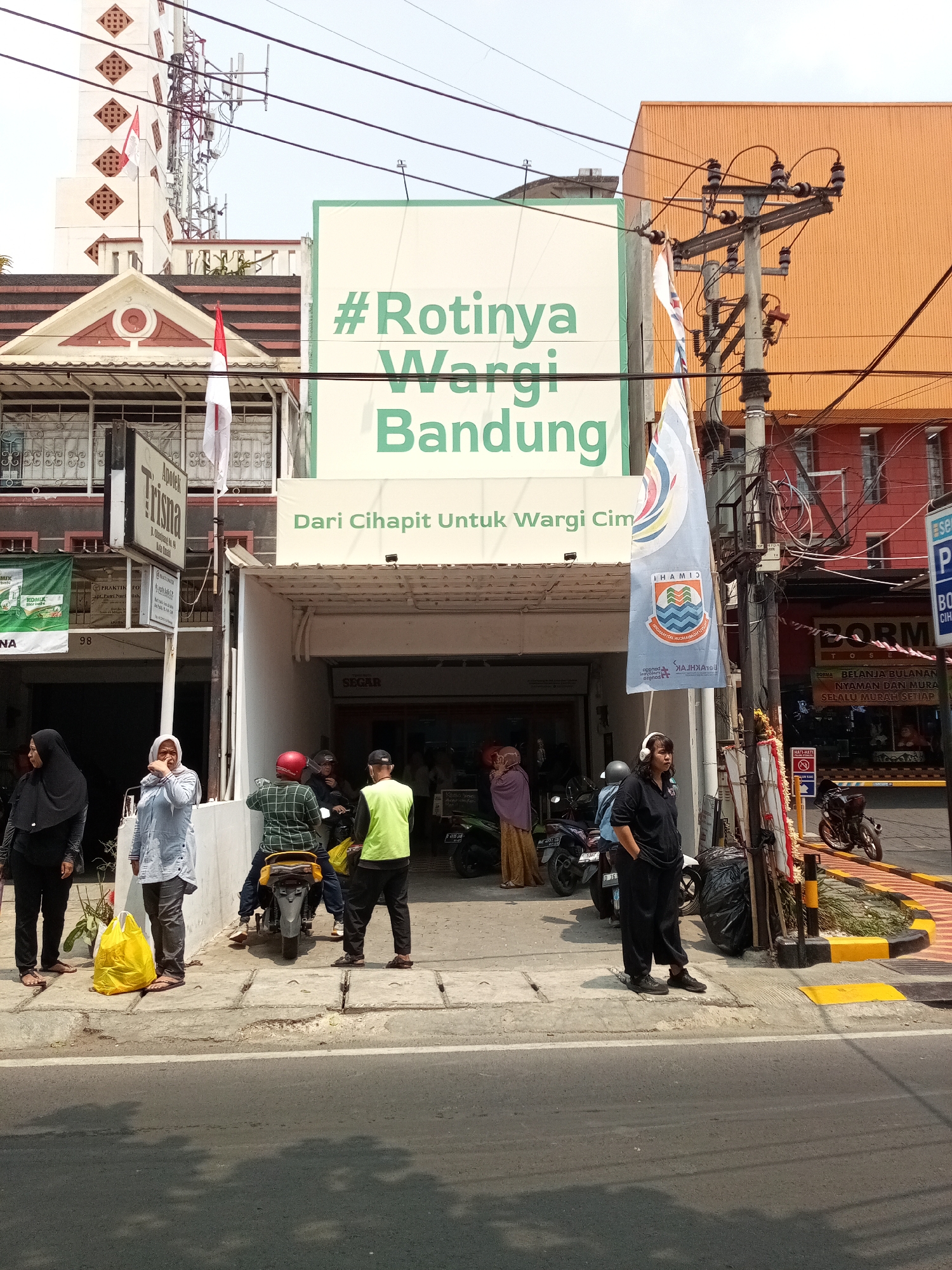 The image depicts a street scene in Cimahi, Indonesia, likely in front of a commercial establishment. A prominent green and white sign reads "#Rotinya Wargi Bandung" and "Dari Cihapit Untuk Wargi Cimahi," indicating a bakery or food business referencing both Bandung and Cimahi. Several people are present on the sidewalk. Two women in hijabs are standing on the left, one in black and the other in light blue. A man wearing a green and black plaid shirt is seated on a motorcycle, facing away from the camera, and another man in a neon green vest is standing to his right. A woman wearing white headphones and a black outfit stands near a trash bin on the right side of the sidewalk. Motorcycles are parked along the sidewalk. The street is paved, with a sidewalk made of concrete slabs. Electrical poles and wires are visible overhead, along with buildings on either side of the street. One building has an orange facade, and another features a white and red architectural style. Indonesian flags are displayed on poles attached to some of the buildings. The environment suggests a sunny day.