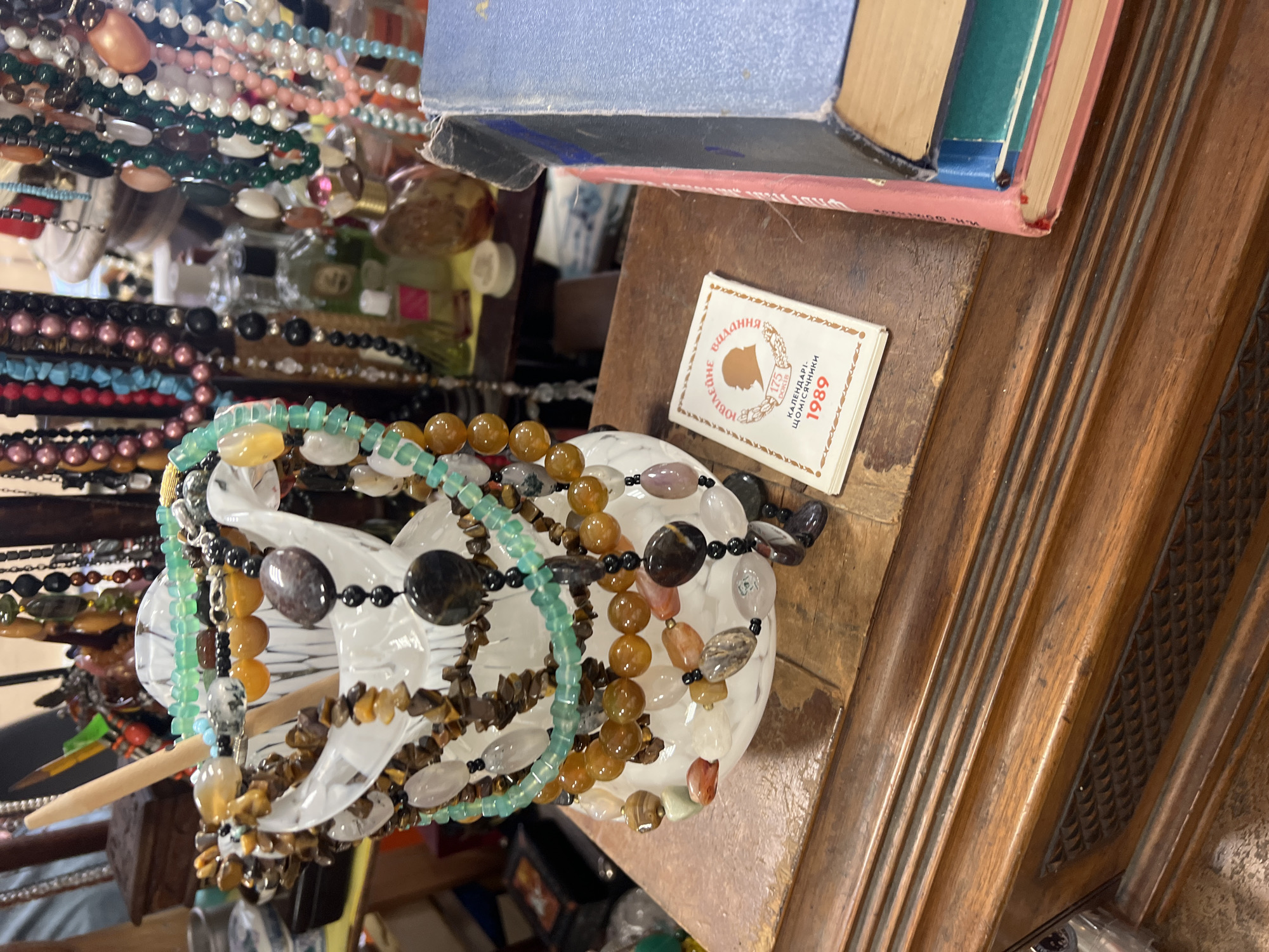 The image depicts a close-up view of a wooden table with various objects on it. In the foreground, to the left, a clear glass vase is filled with an assortment of beaded necklaces. The beads vary in size, color, and material, including what appear to be semi-precious stones like tiger's eye, agate, and jade. A small wooden stick is inserted into the vase among the necklaces. To the right of the vase, a small, rectangular booklet with a light-colored cover is placed on the table. The booklet has text and an illustration of a silhouette of a head. The text appears to be in Cyrillic and includes the year "1989". Next to the booklet, a stack of books is visible, with the top book being blue and the one below it having a pink spine. The background shows a blur of more necklaces and bottles, suggesting a display or collection of items. The table itself is made of dark wood with carved decorative elements. The scene does not provide any explicit indicators of a specific location within Saint Petersburg, Russia, nor does it show any people or specific actions.